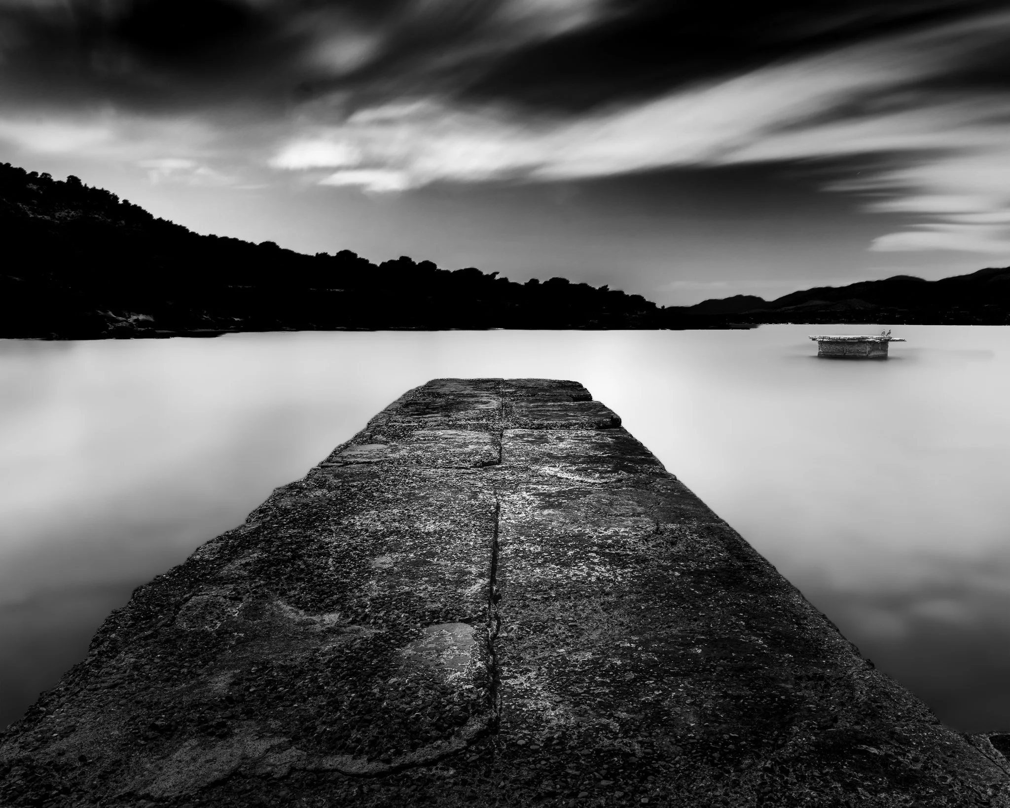 Long exposure black-and-white photo of a pier extending into a calm lake with hills in the background and dramatic cloud streaks in the sky.