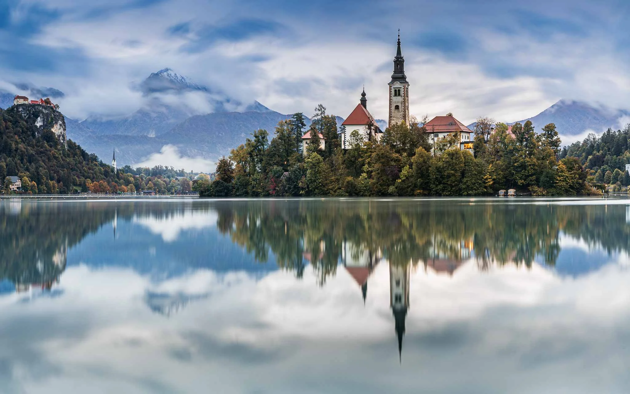 Church on Lake Bled island reflected in calm water beneath cloud-covered Julian Alps in Slovenia.