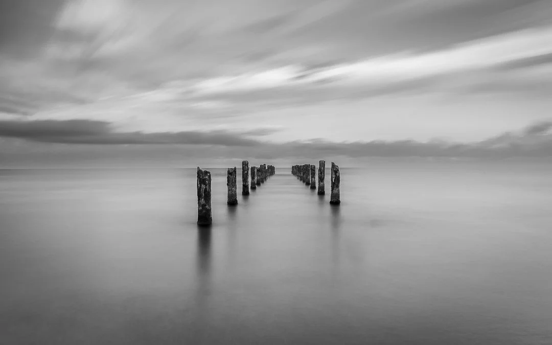 Black and white photo of old wooden posts extending into calm water with a cloudy sky overhead.