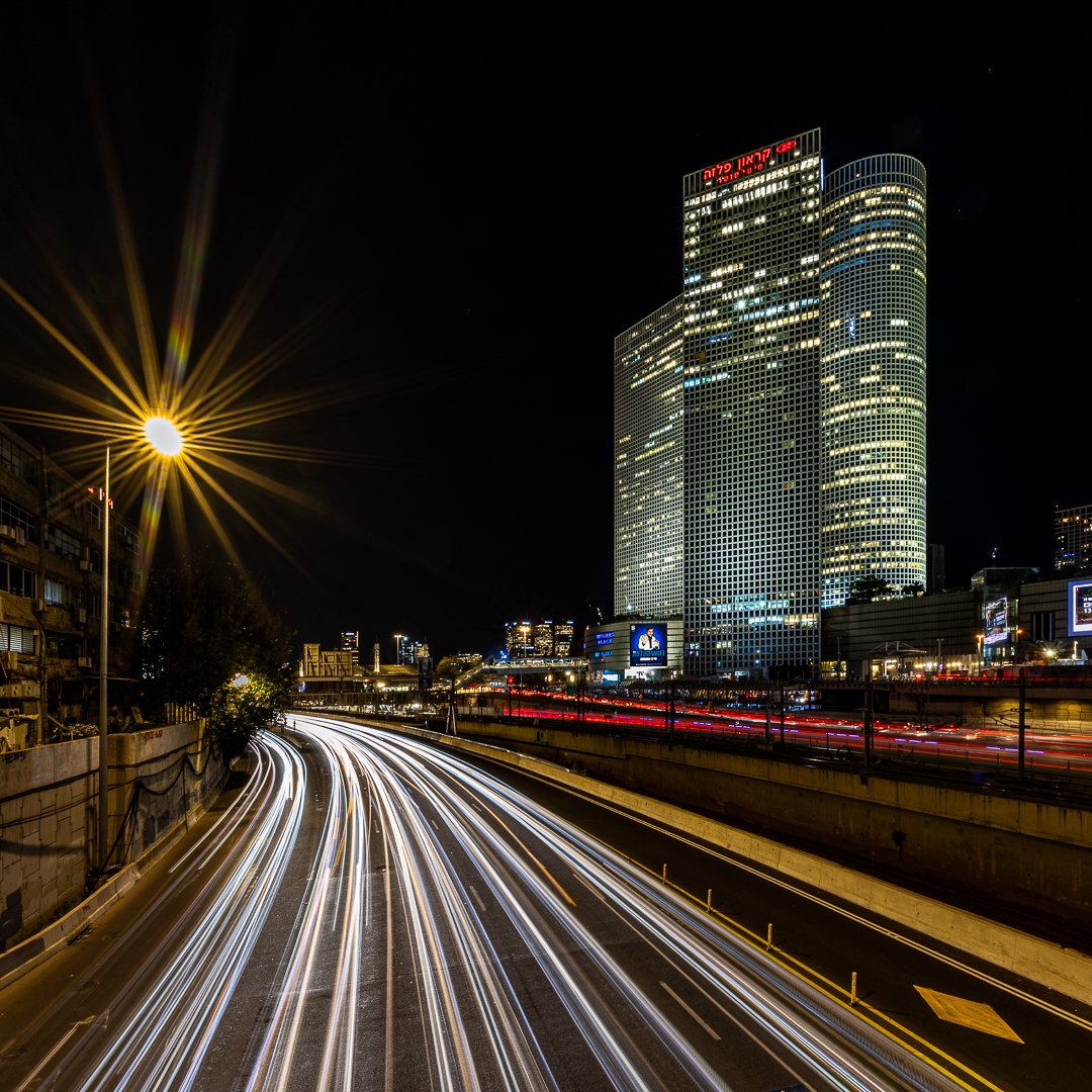Nighttime cityscape with light streaks from moving vehicles, tall office buildings with illuminated windows, and a bright streetlight creating a starburst effect.