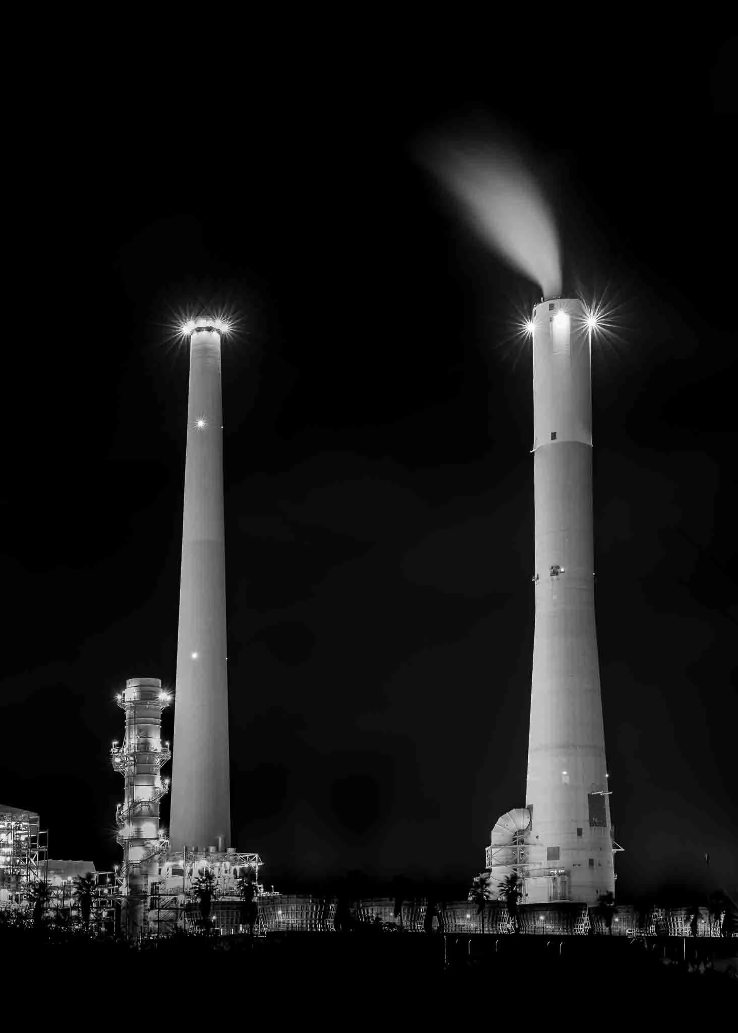 lack-and-white night photograph of two illuminated industrial smokestacks, with steam rising from one tower against a dark sky.