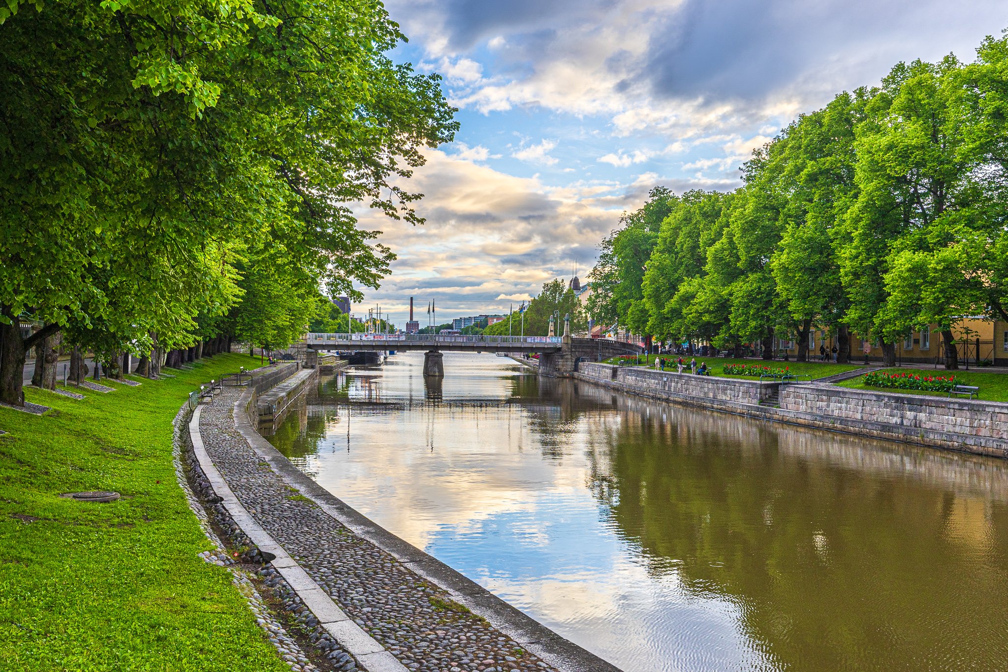 A scenic view of a river lined with green trees on both sides, with a bridge in the distance. The sky is partly cloudy with patches of blue. People are walking or sitting along the riverbank, and colorful flowers are planted beside the path.