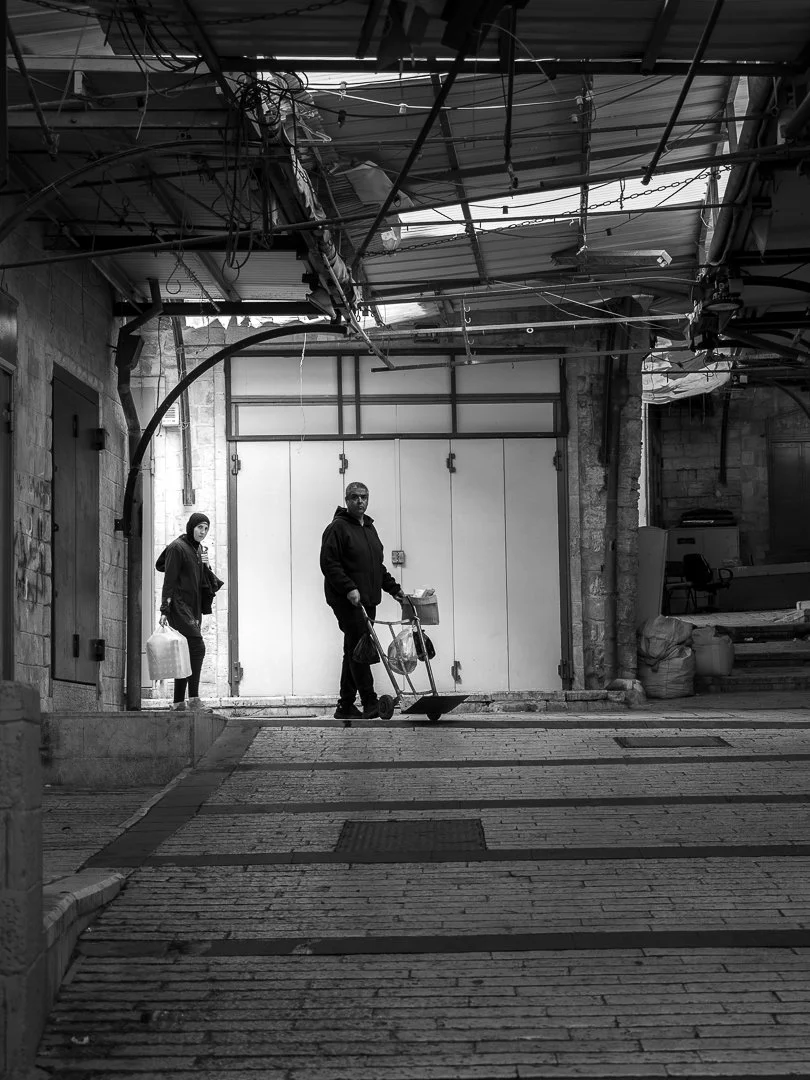 Two people standing in an urban alleyway. One person, wearing a dark hoodie and pants, is pushing a hand trolley with bags. The other person, dressed in a jacket and head covering, is carrying a bag and standing near a wall. The alley has an overhead