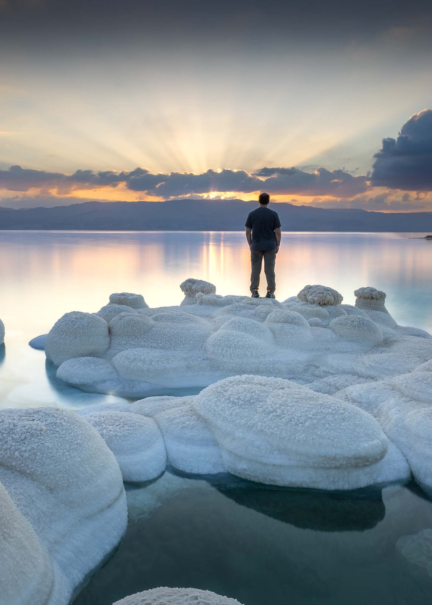 A lone figure stands among salt formations at the Dead Sea as dramatic sun rays break through clouds over the mountains.