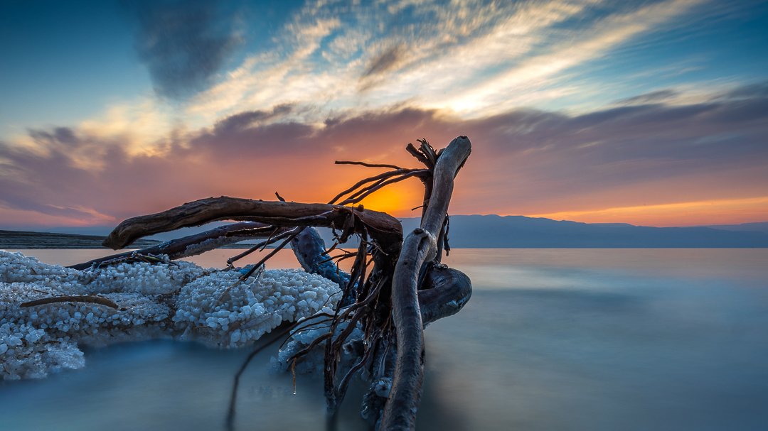 A driftwood log lying on salt deposits along a tranquil shoreline during sunset, with a colorful sky and distant mountains in the background.