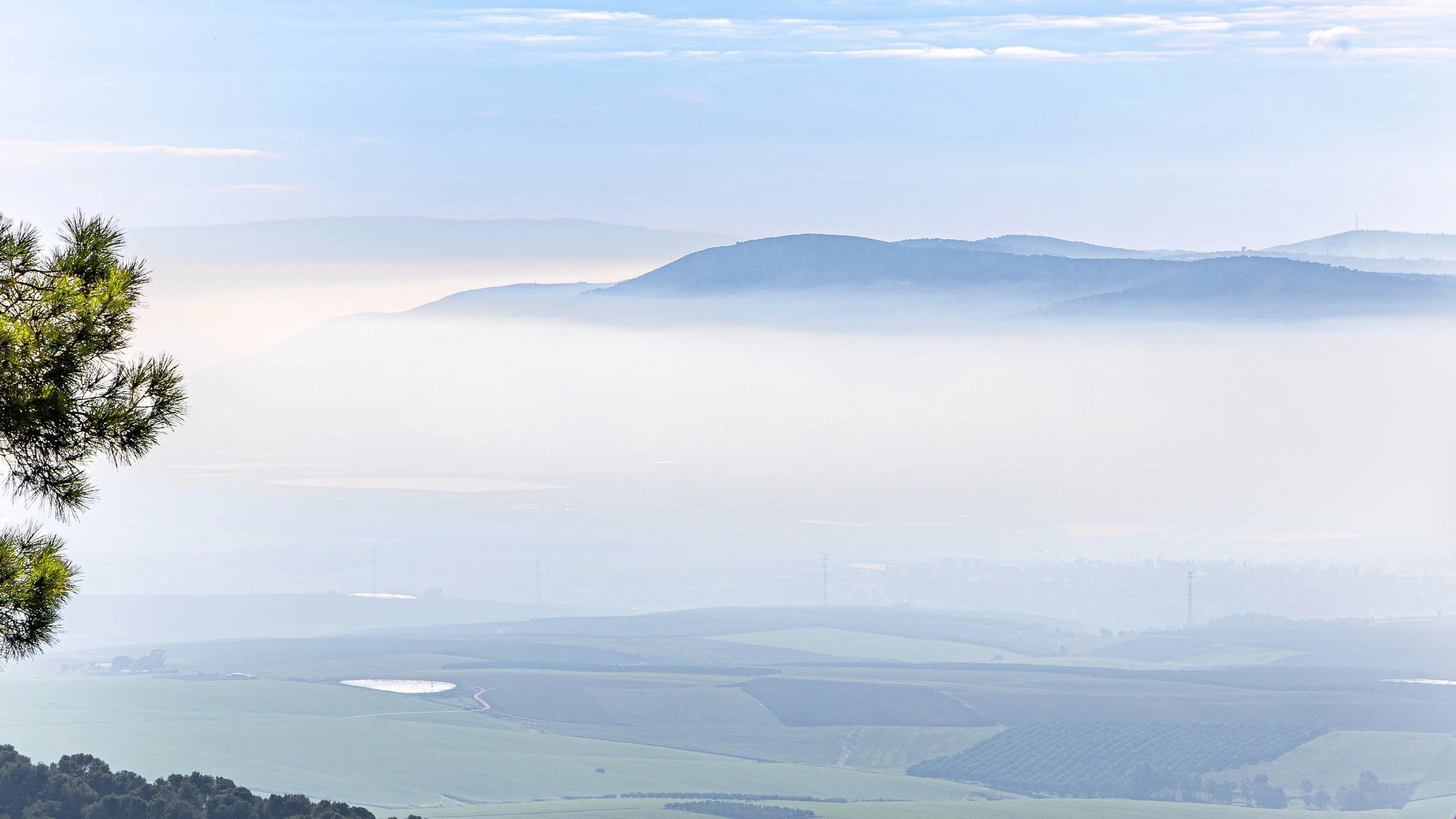 A landscape view of distant mountains shrouded in mist, with a clear blue sky and a tree on the left side of the image.