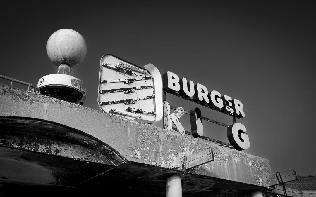 Black and white photo of an old, weathered fast food restaurant sign that reads "Burger King" with some letters missing and a large spherical light fixture nearby.