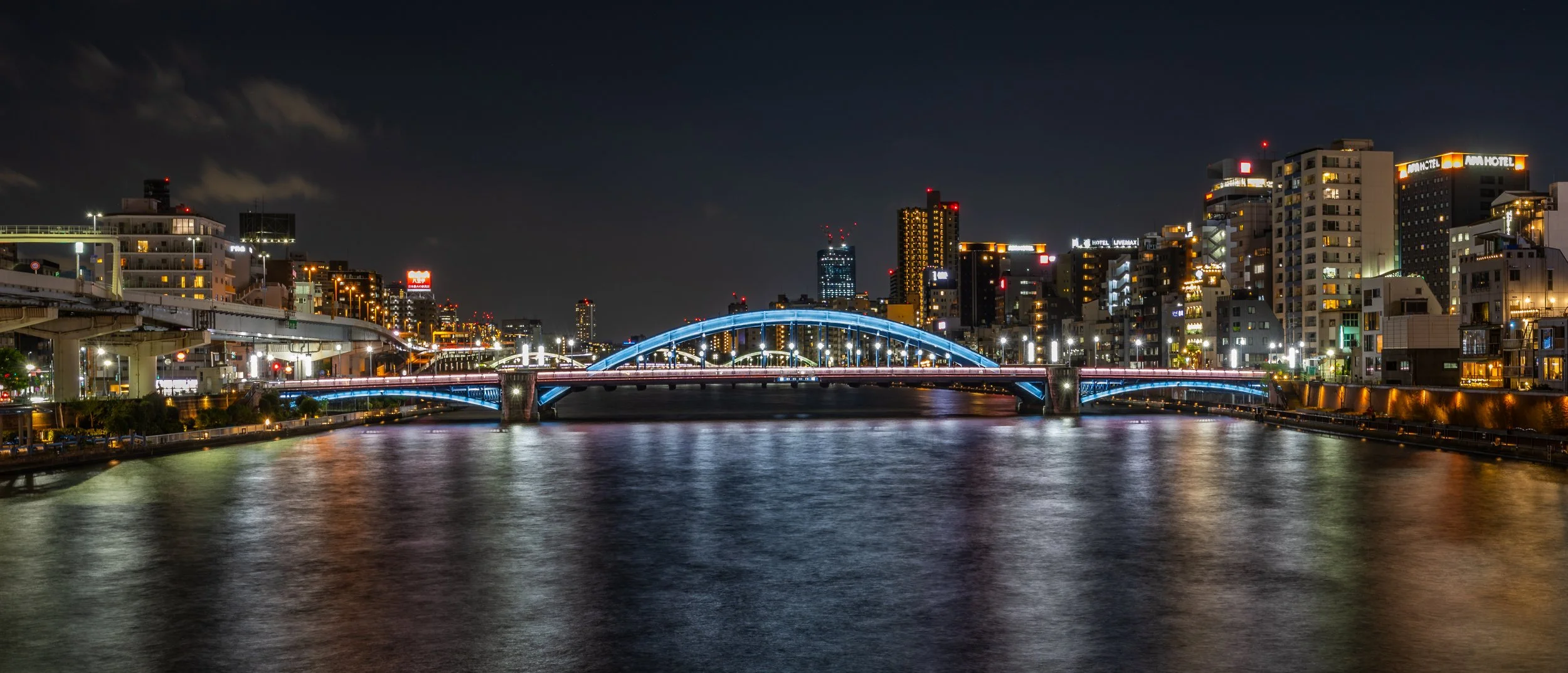 Night view of a city skyline with illuminated buildings and a bridge over a river, reflecting city lights on the water.