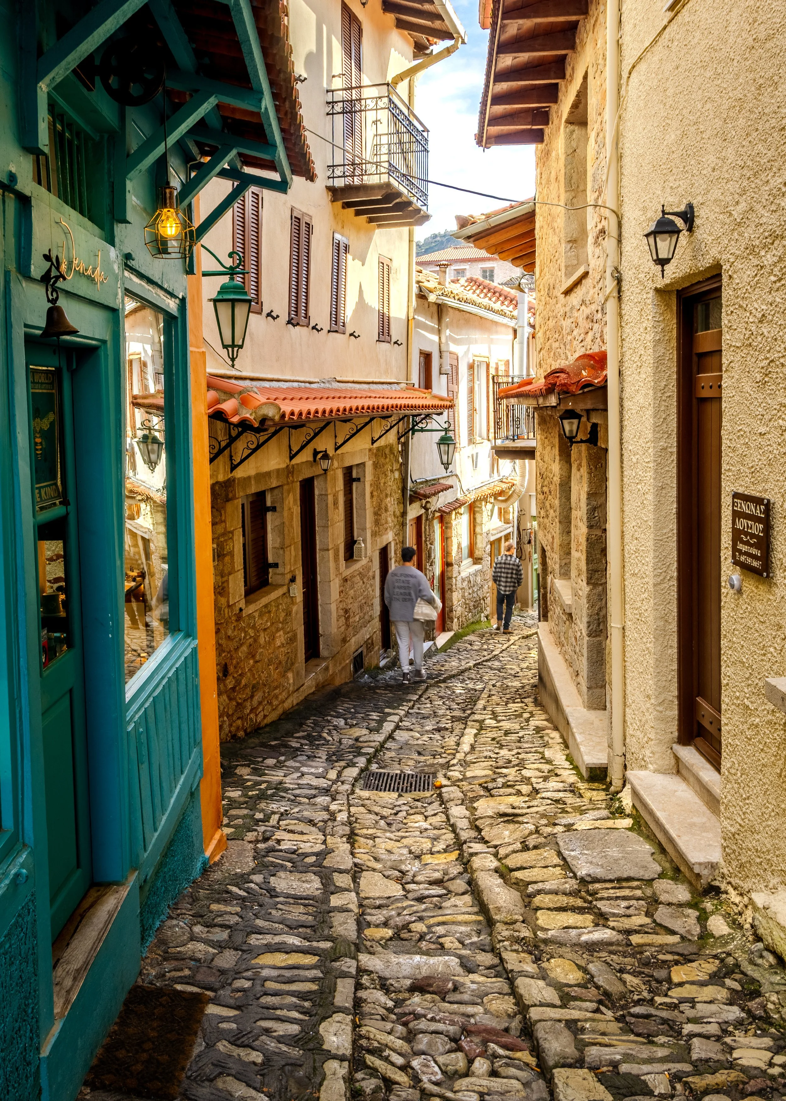 Narrow cobblestone street with stone houses, balconies and two people walking downhill.