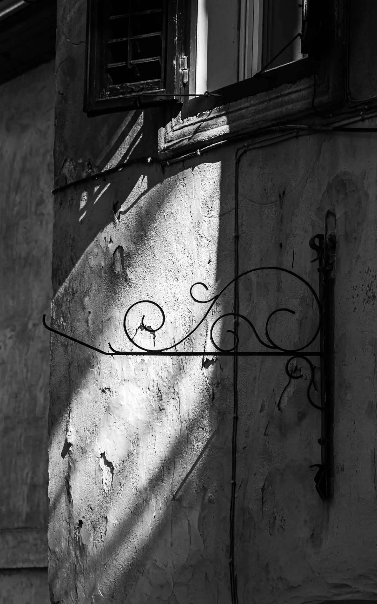 Black and white architectural photograph of an ornate wrought-iron bracket casting curved shadows across a weathered wall, intersected by dramatic diagonal sunlight in Slovenia.