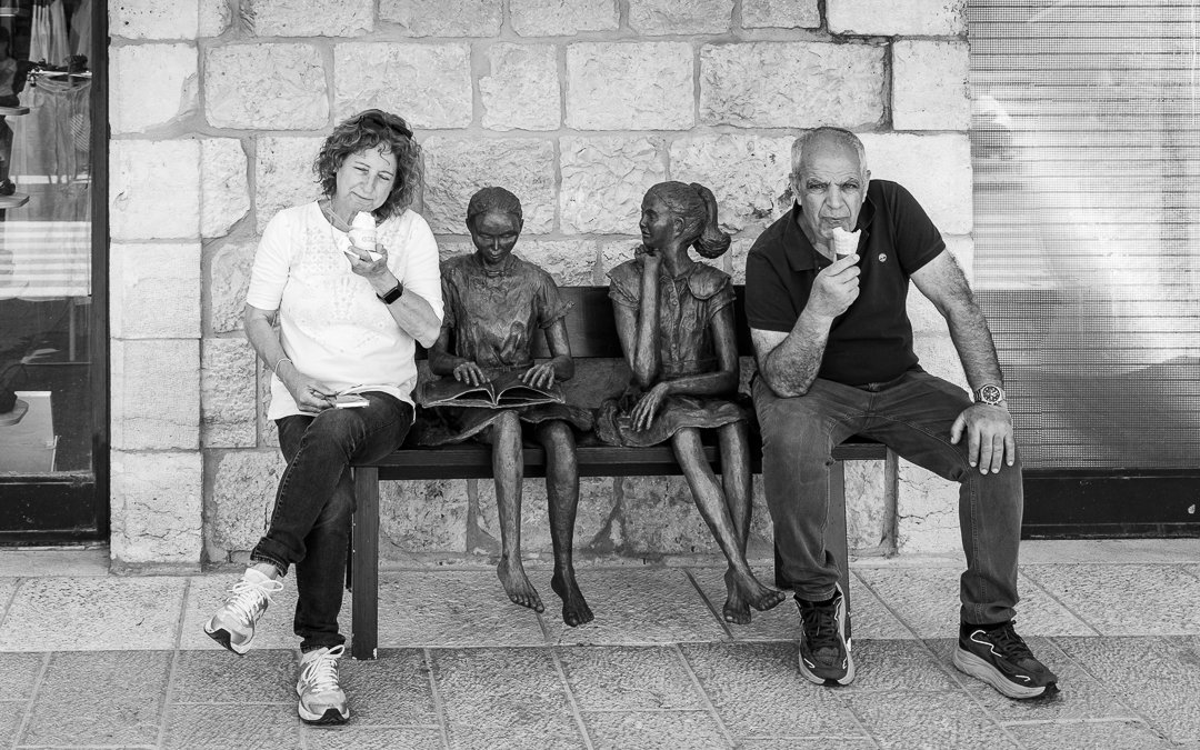 Two adults, a woman and a man, sitting on a bench with two bronze statues of children, all eating ice cream cones.