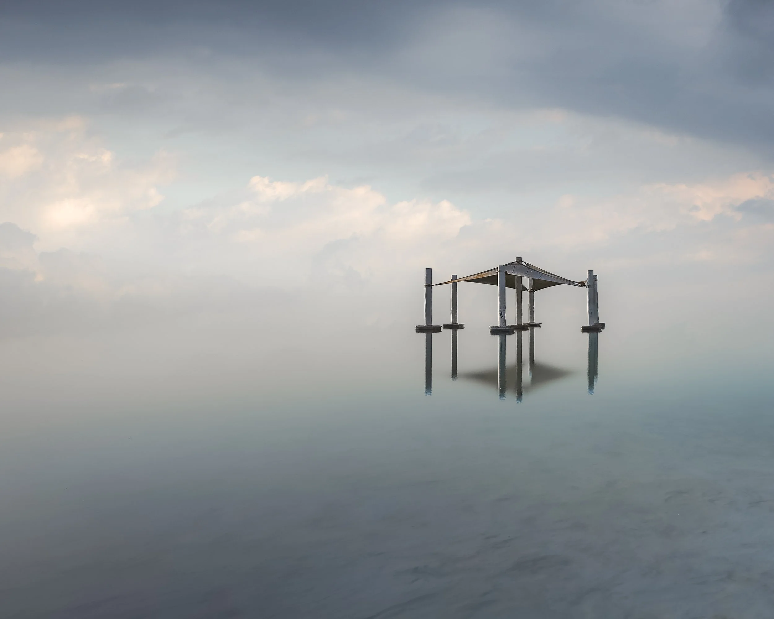 A calm ocean scene with a small wooden hut on stilts at the water's edge, under a cloudy sky.