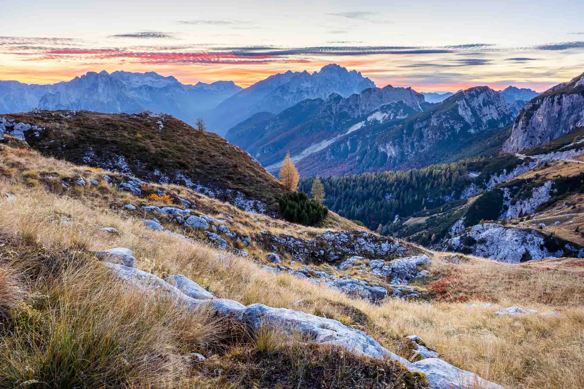 Rocky foreground and golden alpine grass overlooking layered mountain ridges beneath a colorful sky near Mangart Saddle, Slovenia.