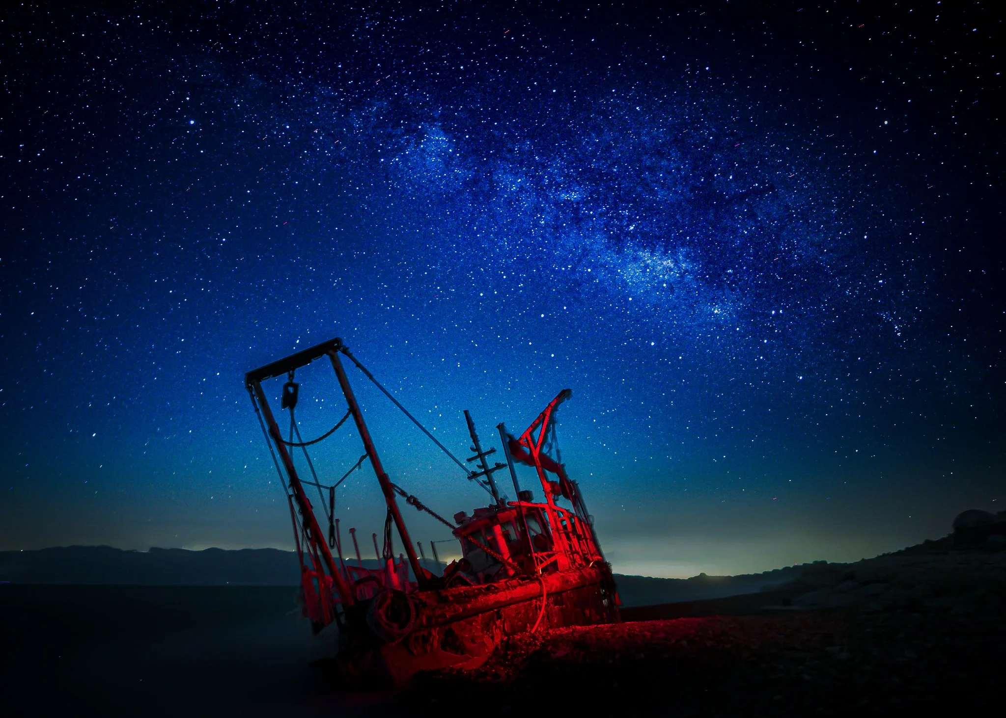 An old red boat on the shore under a star-filled night sky, with the Milky Way visible.