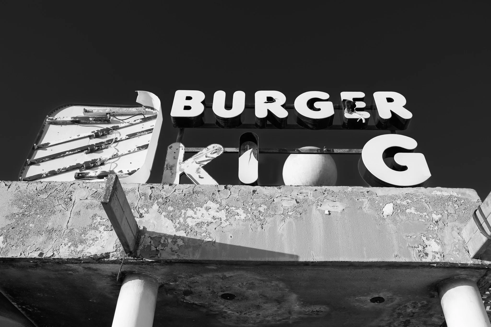 Black and white photo of an old, worn sign reading "BURGER KING" with some letters missing, mounted on a weathered building rooftop.