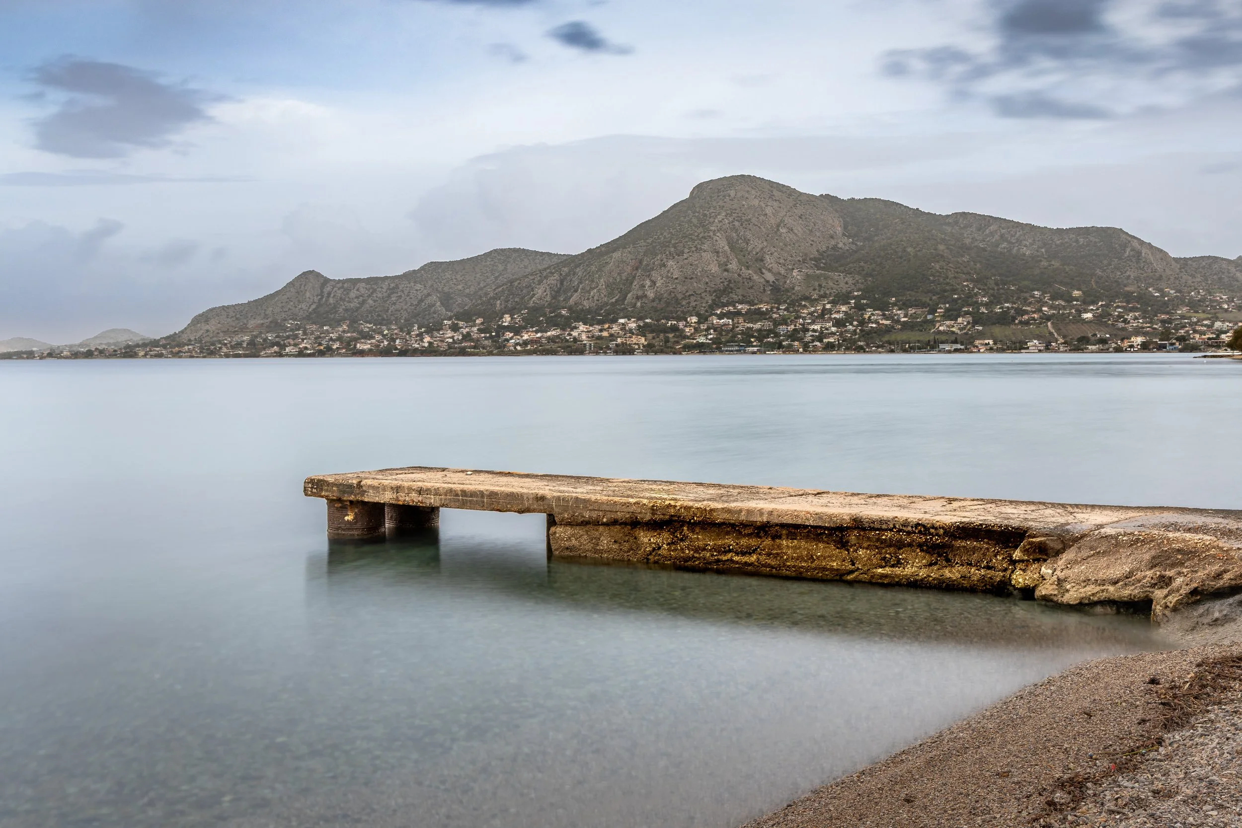 A peaceful lakeside scene with a weathered wooden dock extending into calm water, with mountains and a cloudy sky in the background, photograph by erez nudmanov