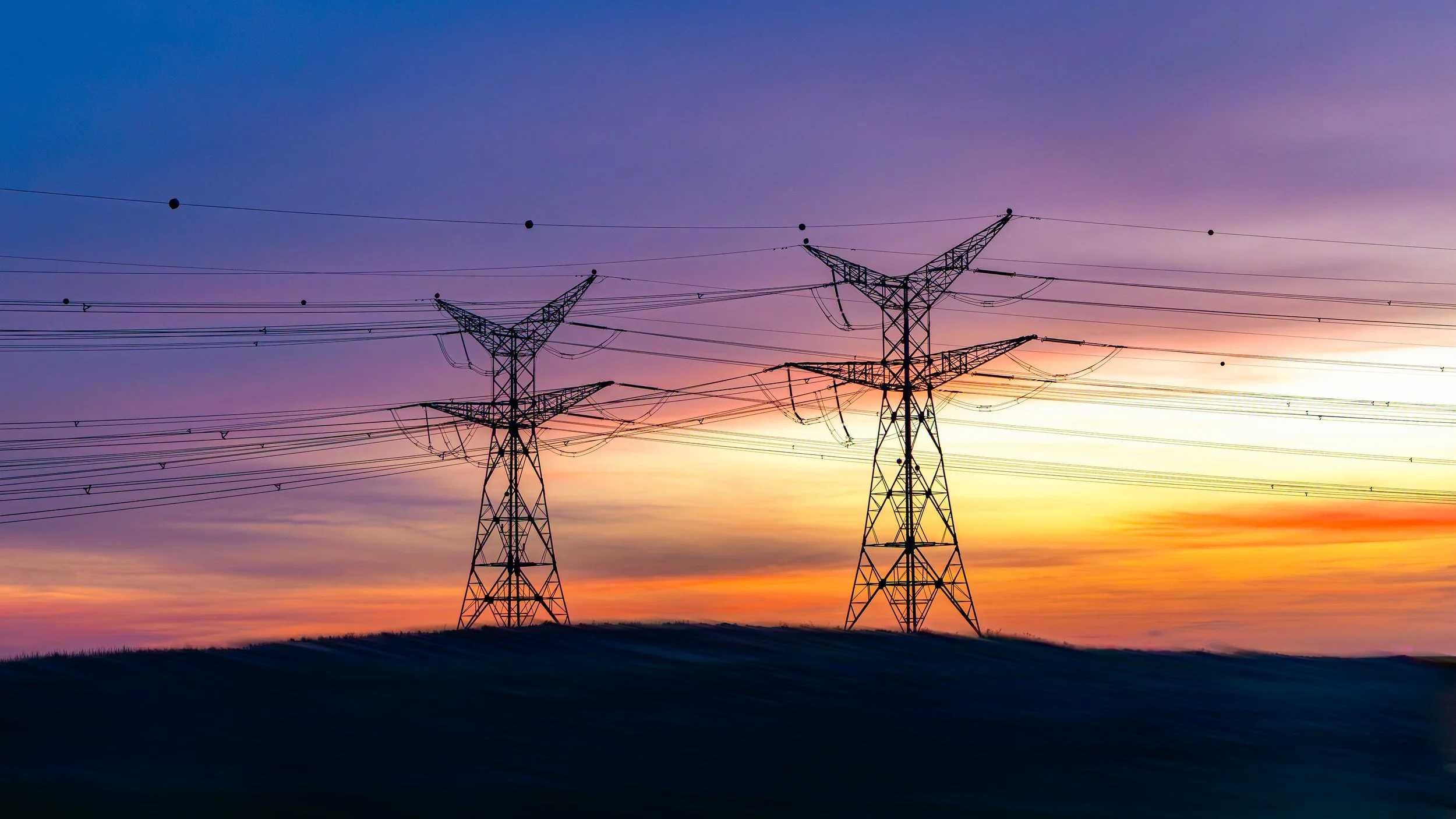 Silhouettes of power transmission towers against a colorful sunset sky, with a rural landscape in the foreground.