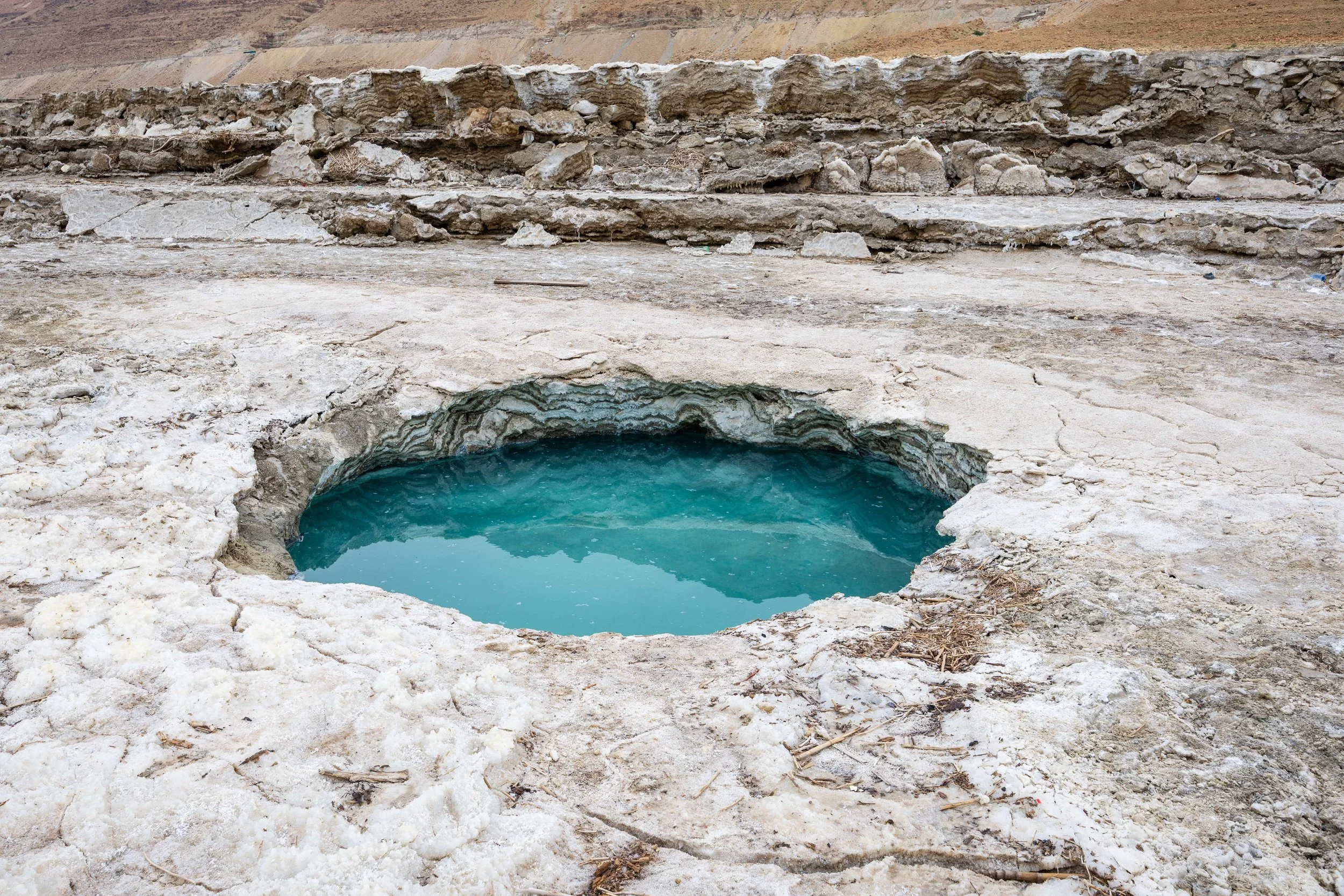 A hot spring with blue water surrounded by white mineral deposits in a barren, rocky landscape.
