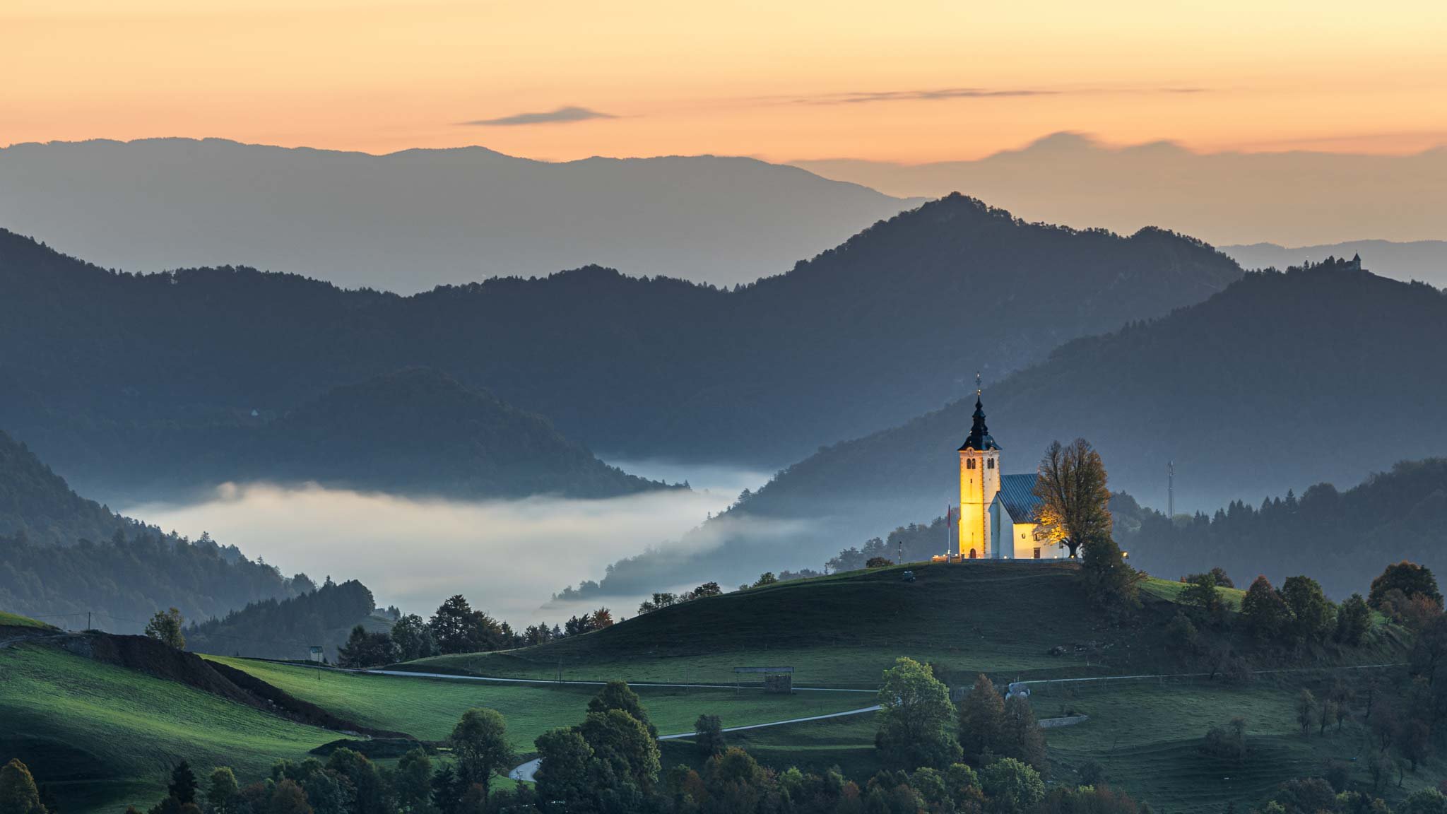 Illuminated Jamnik church on a grassy ridge at sunrise, with layered mountains beneath an orange autumn sky in Slovenia.