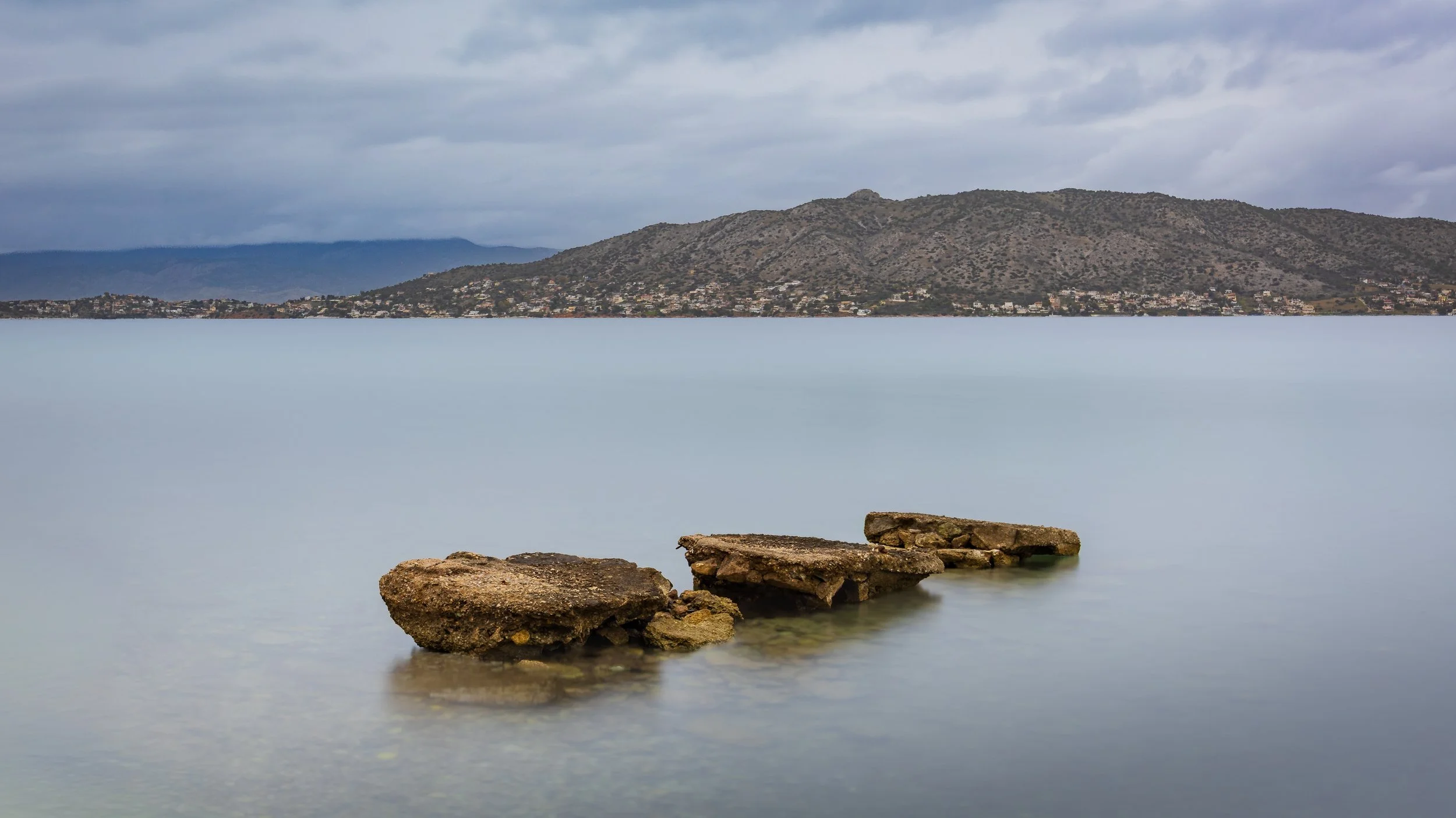 Four rocks partially submerged in a calm lake with mountains and a cloudy sky in the background, photograph by erez nudmanov