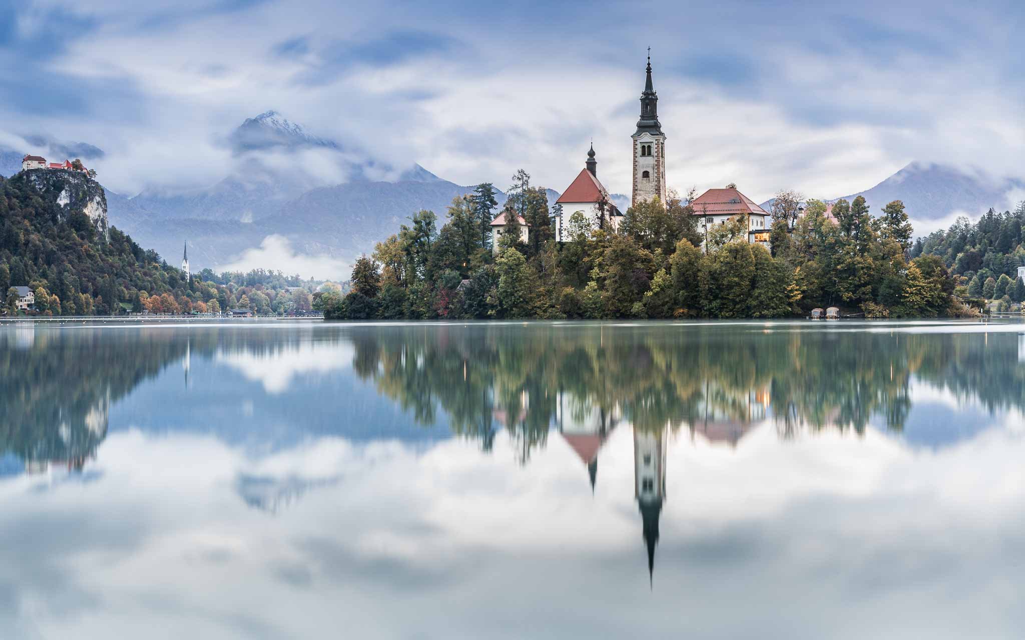 Lake Bled island church and Bled Castle reflected in calm water, with autumn foliage and mist covered mountains in Slovenia.