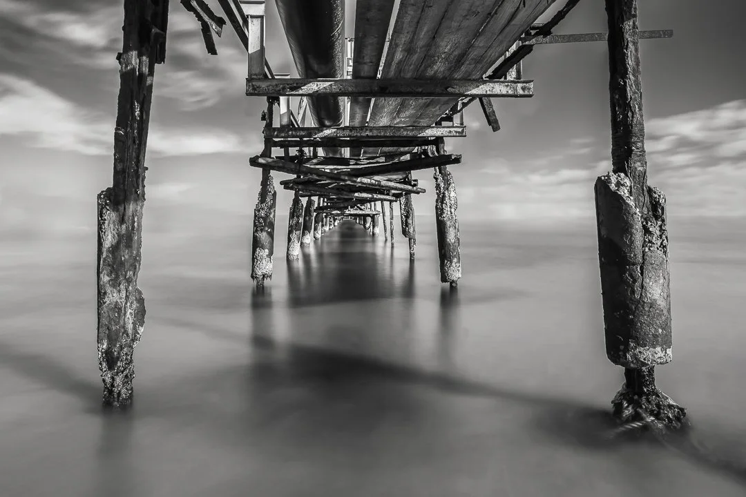 Underwater view of a weathered wooden pier with barnacle-covered supports extending into calm water, captured in black and white.