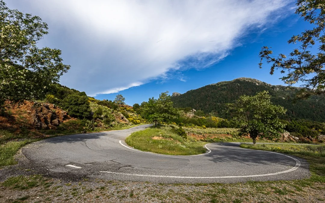 A winding mountain road surrounded by green trees and rocks under a partly cloudy sky.