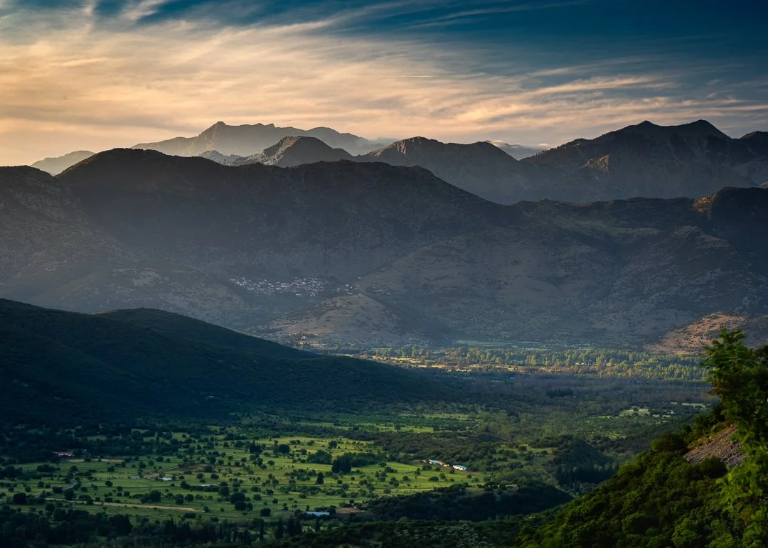 Scenic view of a valley with green fields and scattered trees, surrounded by mountain ranges under a partly cloudy sky during sunset.
