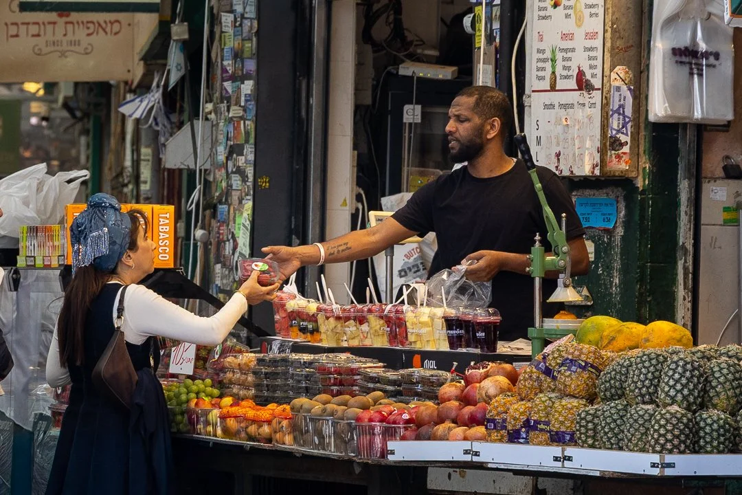 A man selling fruit and desserts at an outdoor market, handing a container to a woman shopper.