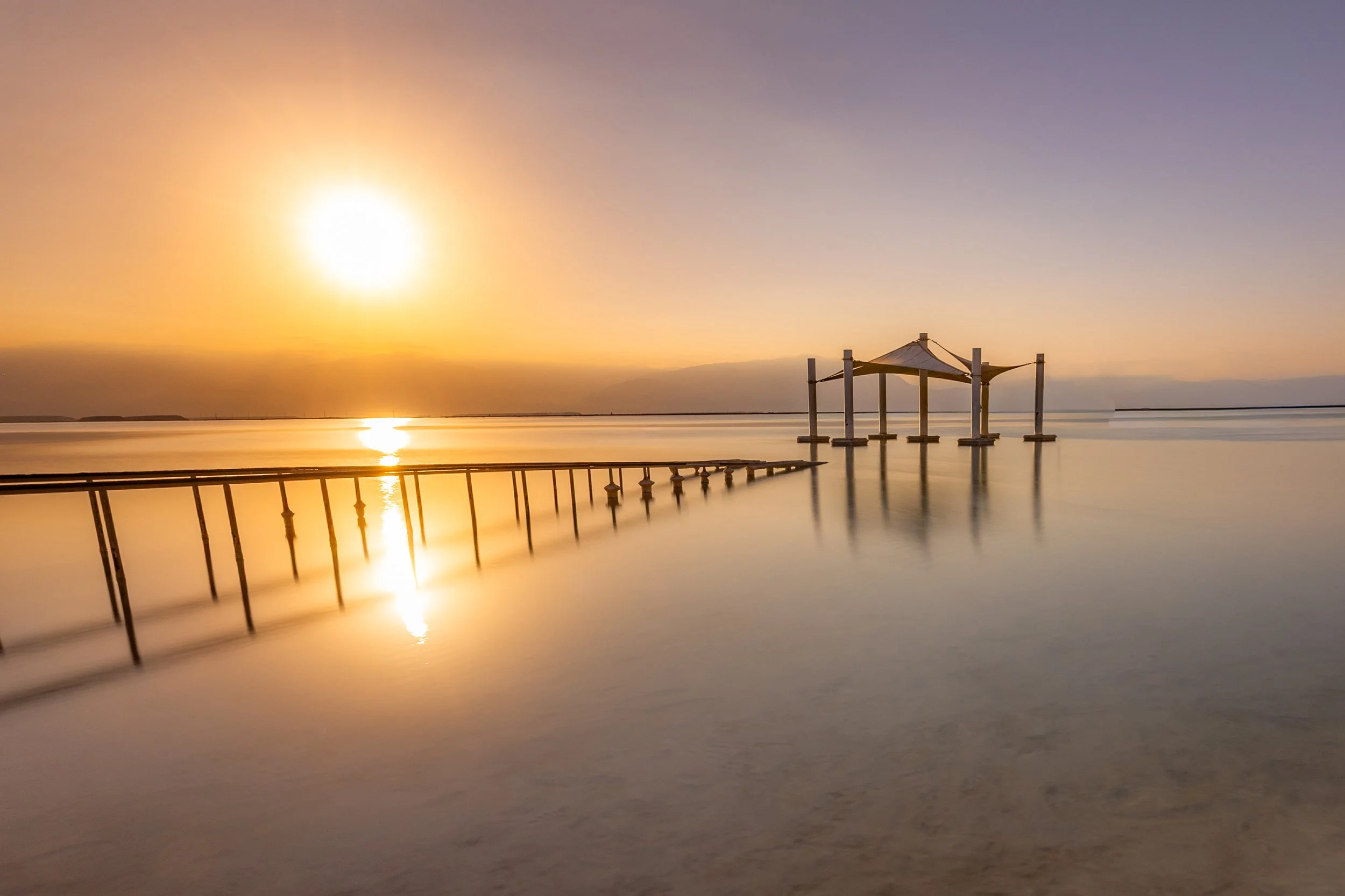 Sunset over calm water with a pier leading to a shaded gazebo, reflecting on the surface, under a colorful sky.