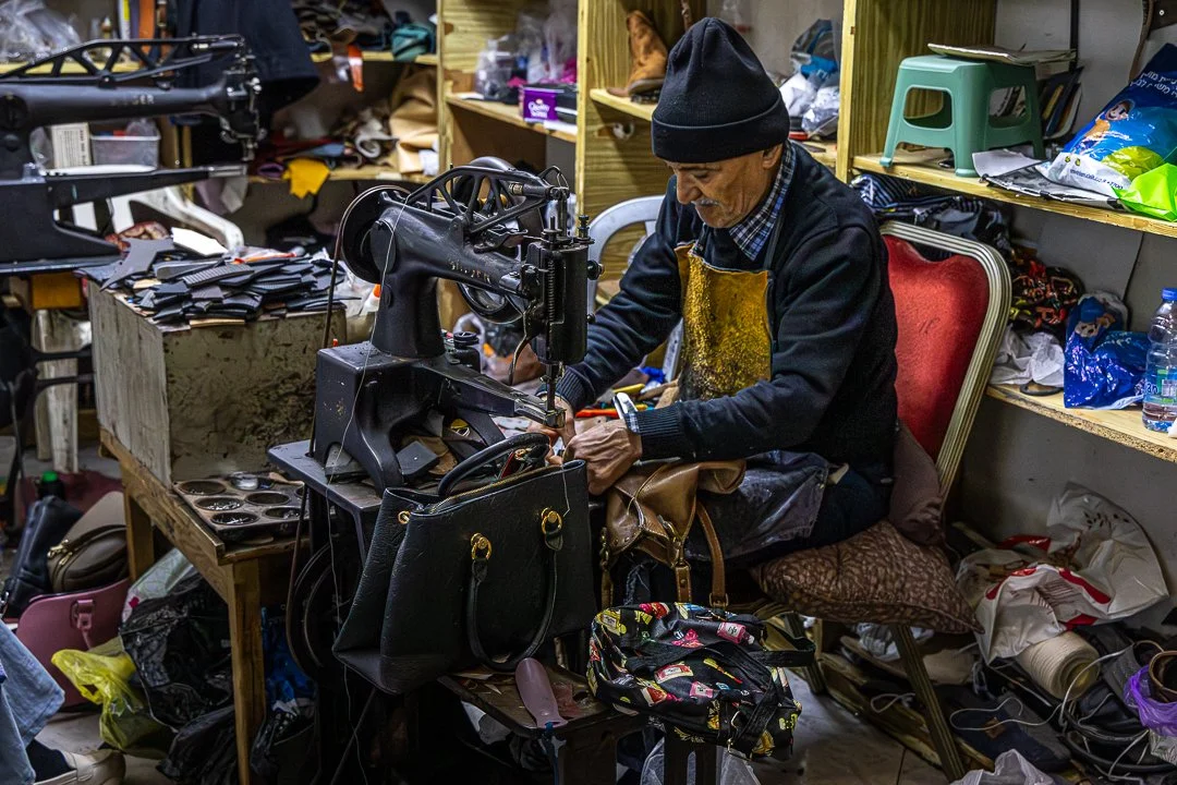 An elderly man working with a sewing machine amid clutter in a small workshop. Shelves filled with shoes, bags, and supplies surround him.