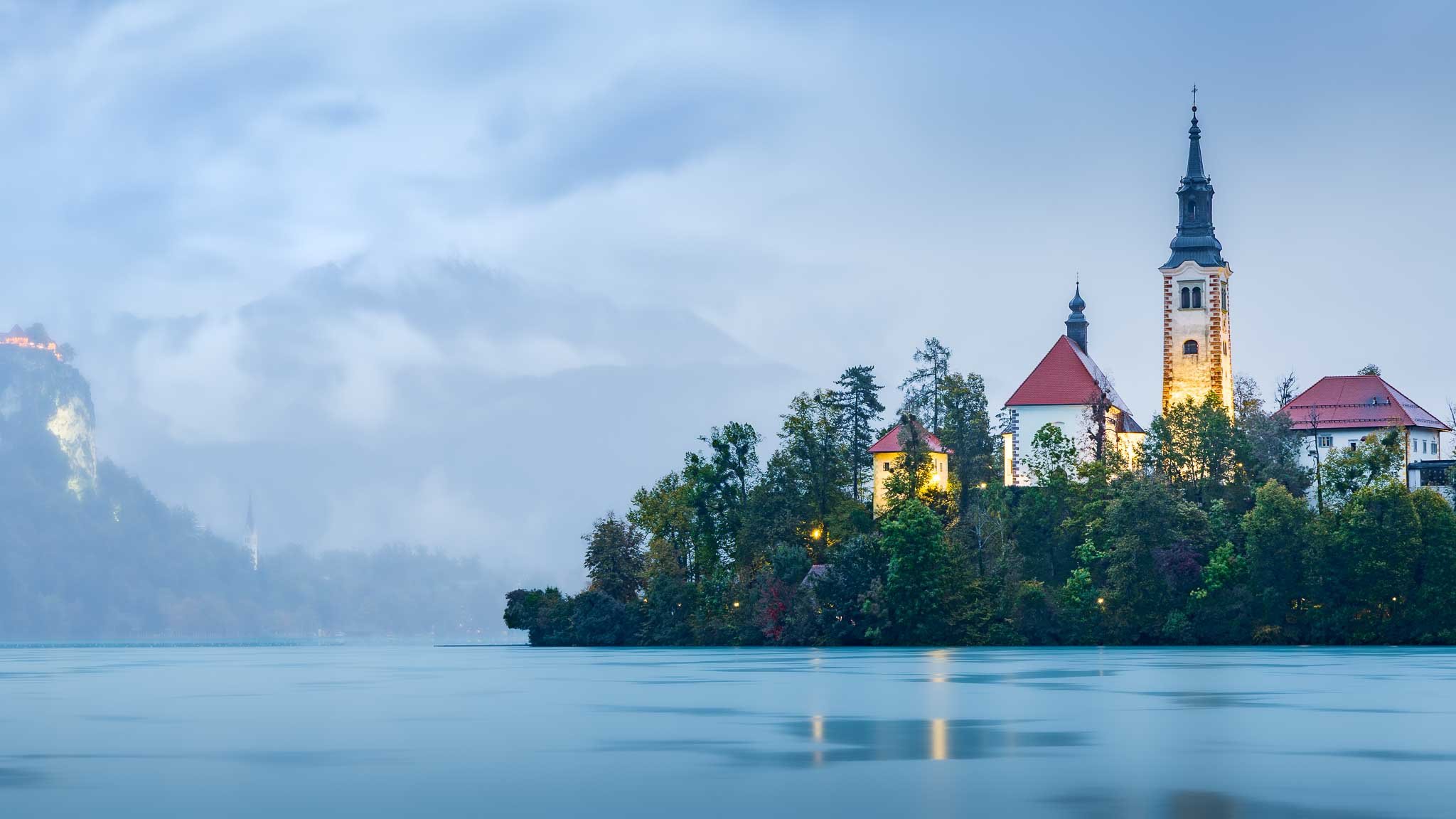 Illuminated church on Lake Bled island emerging from blue-hour mist above smooth lake water in Slovenia.
