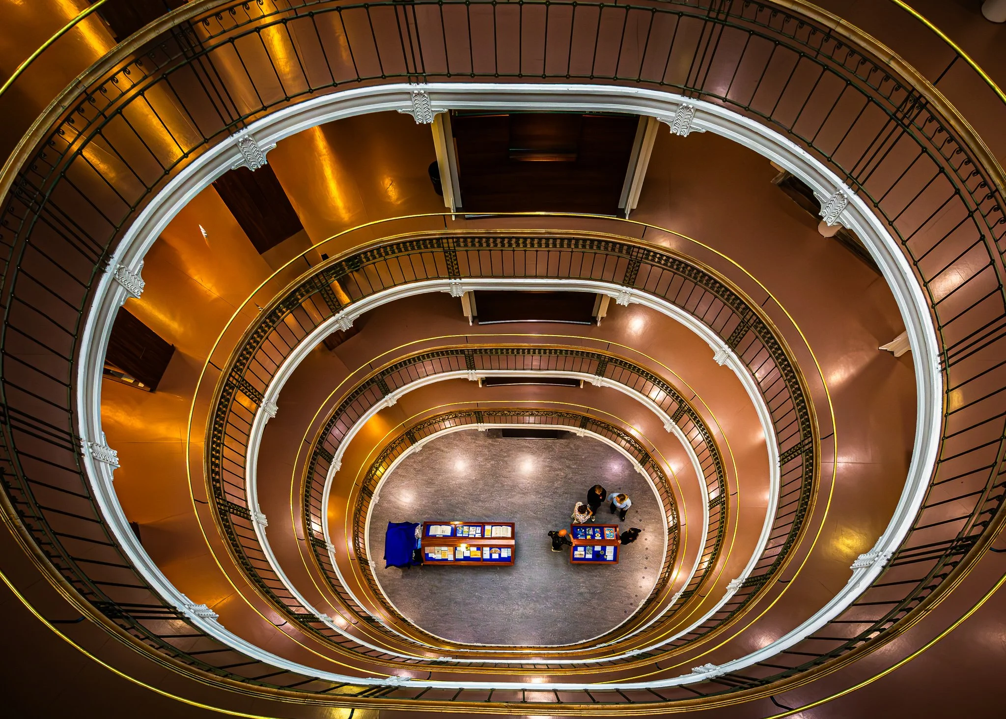 A spiral staircase viewed from above showing golden handrails and white decorative elements. At the bottom, three people are standing near display tables in a gallery or museum setting.