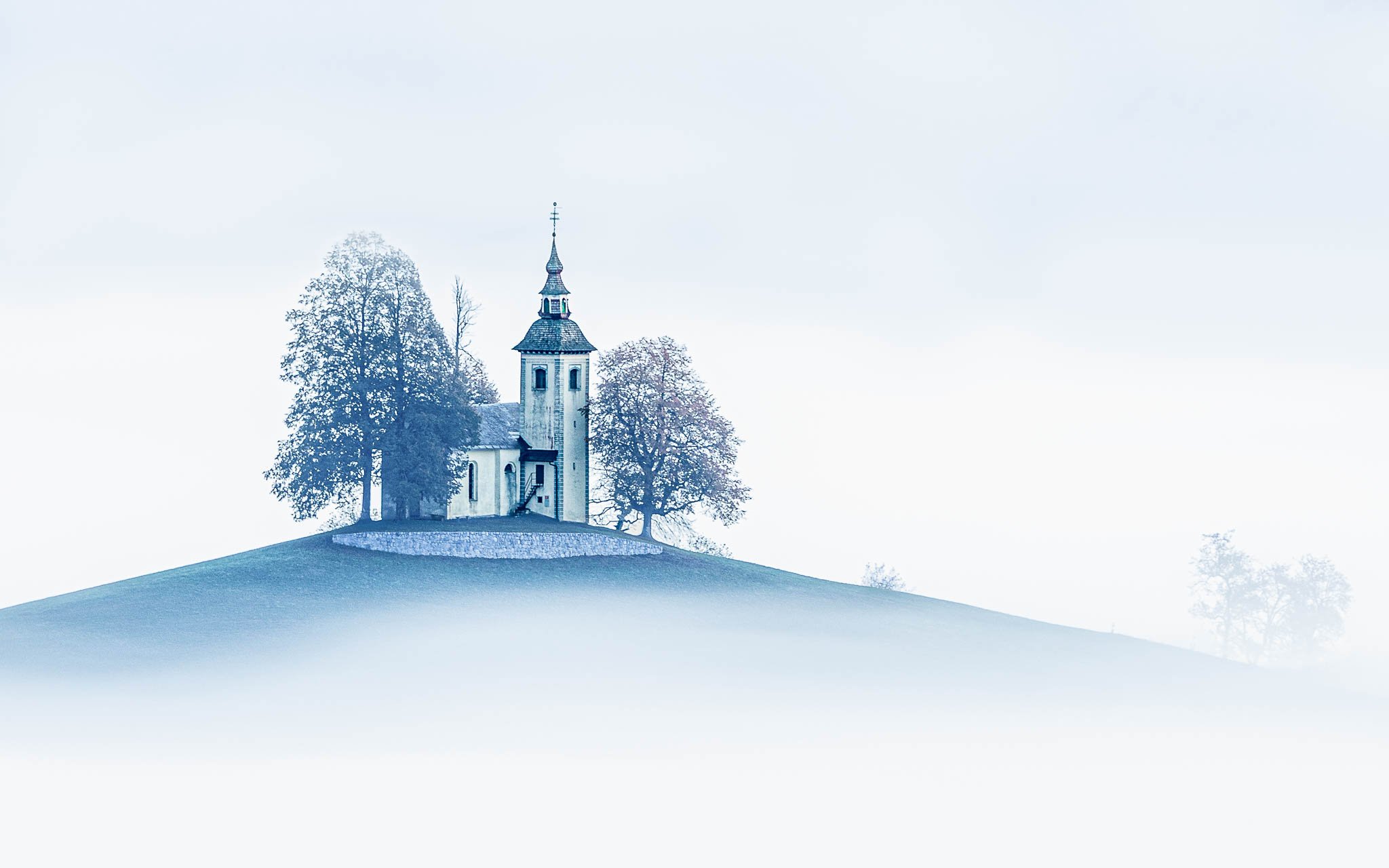 St. Thomas Church and surrounding trees emerging from autumn fog, with soft blue tones and open white space, Slovenia.