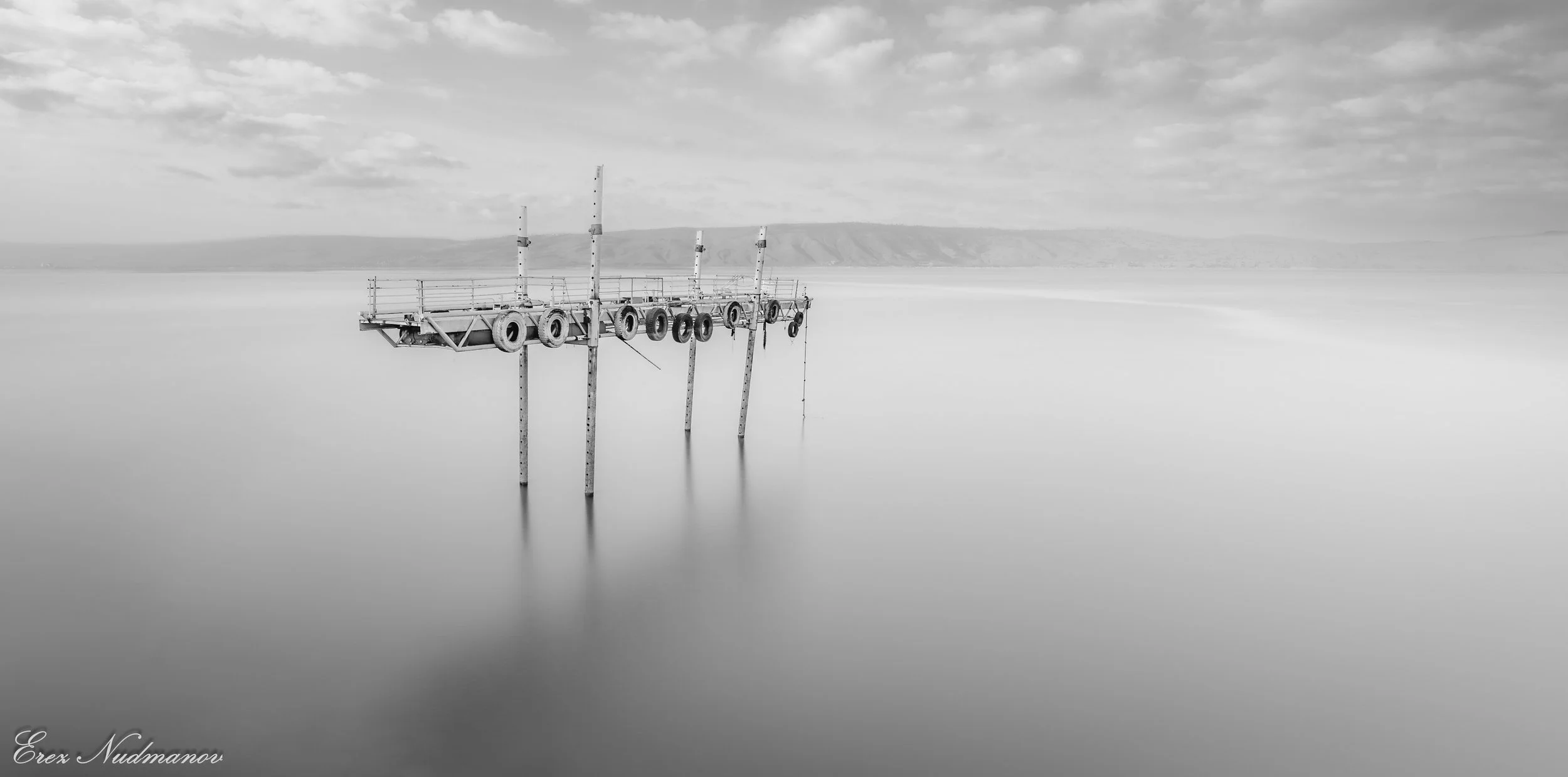 Black and white photo of a partially submerged pier with tires hanging from it, extending into calm water, with hills in the background and a cloudy sky overhead.