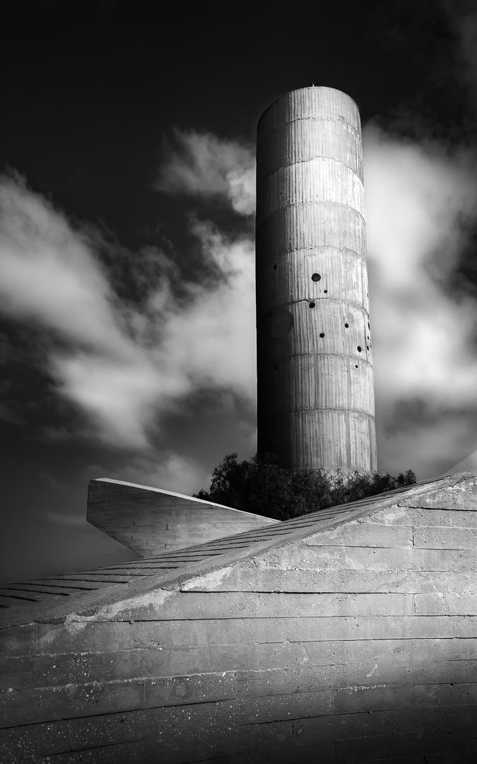 Black and white photograph of a tall, cylindrical concrete tower with evenly spaced circular holes, set against a cloudy sky. In the foreground, there are angled, textured concrete stairs or platform leading up to the tower.