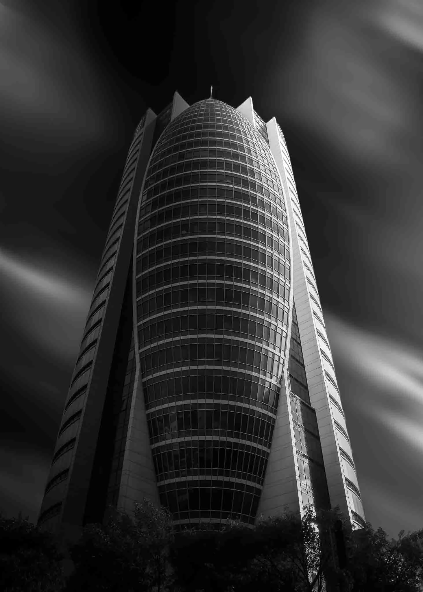 Black and white long-exposure architectural photograph of Haifa’s Sail Tower, rising dramatically against streaked clouds.