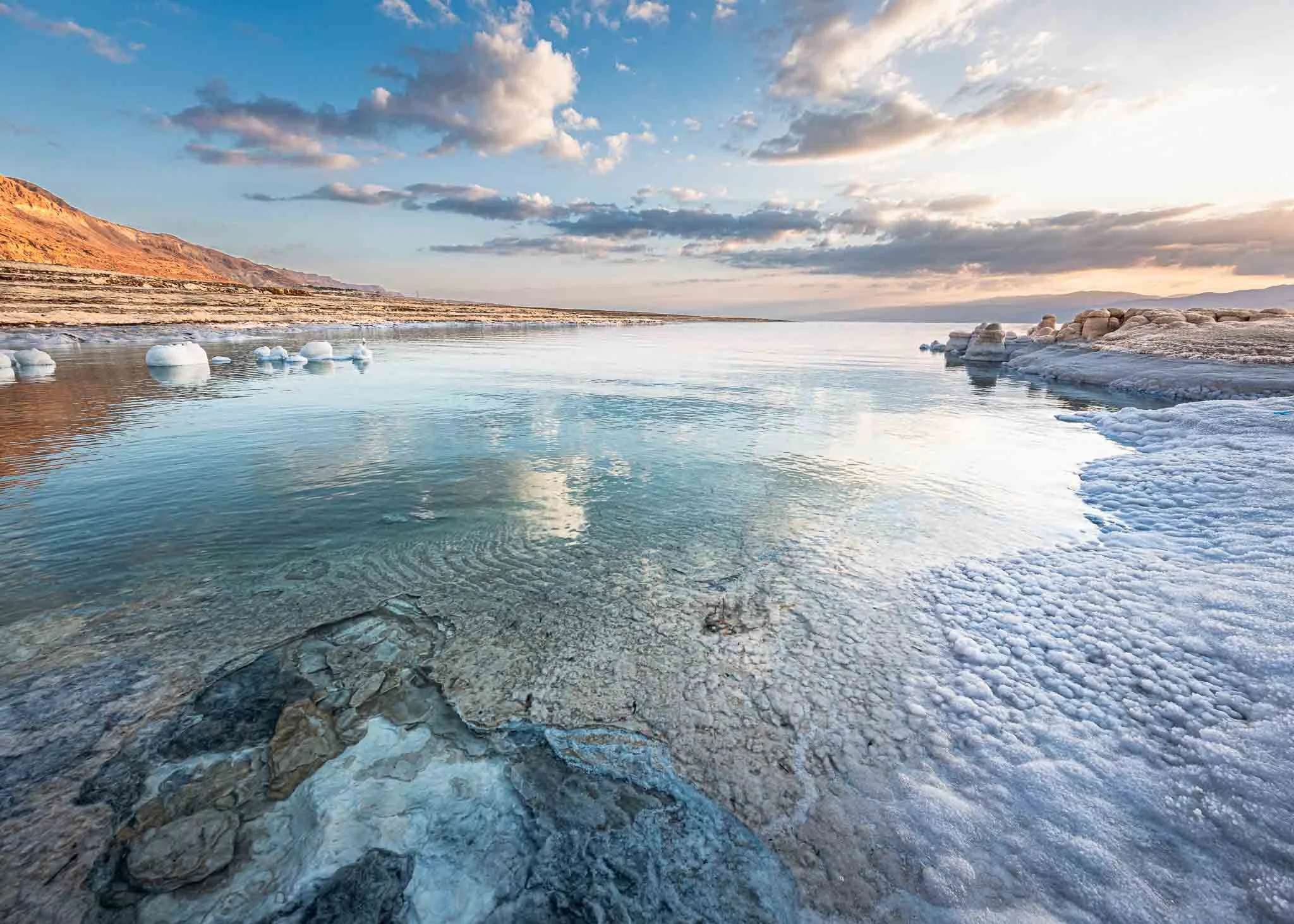 Salt formations and shallow water at the Dead Sea reflect soft evening light beneath a dramatic cloud-filled sky.
