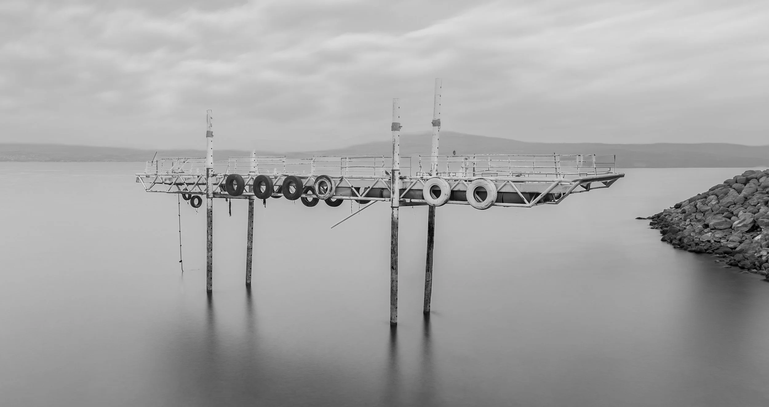 A weathered, unused metal pier with tires hanging along its side, extending over calm water. The sky is overcast and there is a rocky shoreline on the right.