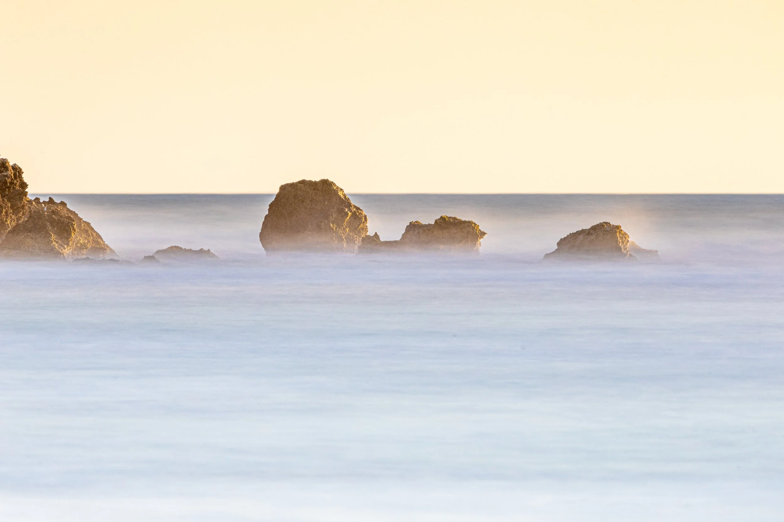 Seascape with large rocks and smooth water under a light-colored sky.