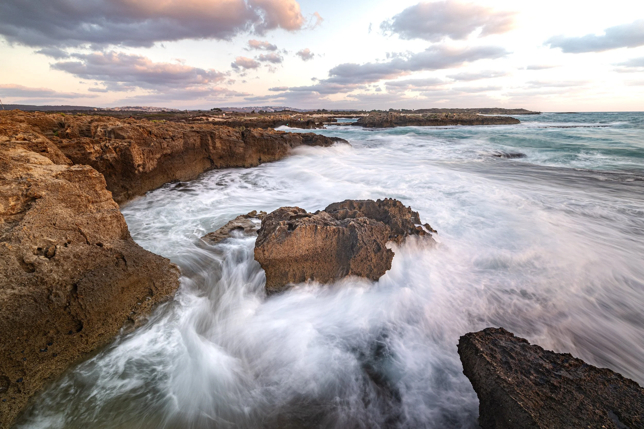 Coastal scene with crashing waves on rocky shoreline during sunset, cloudy sky, and distant land on horizon.