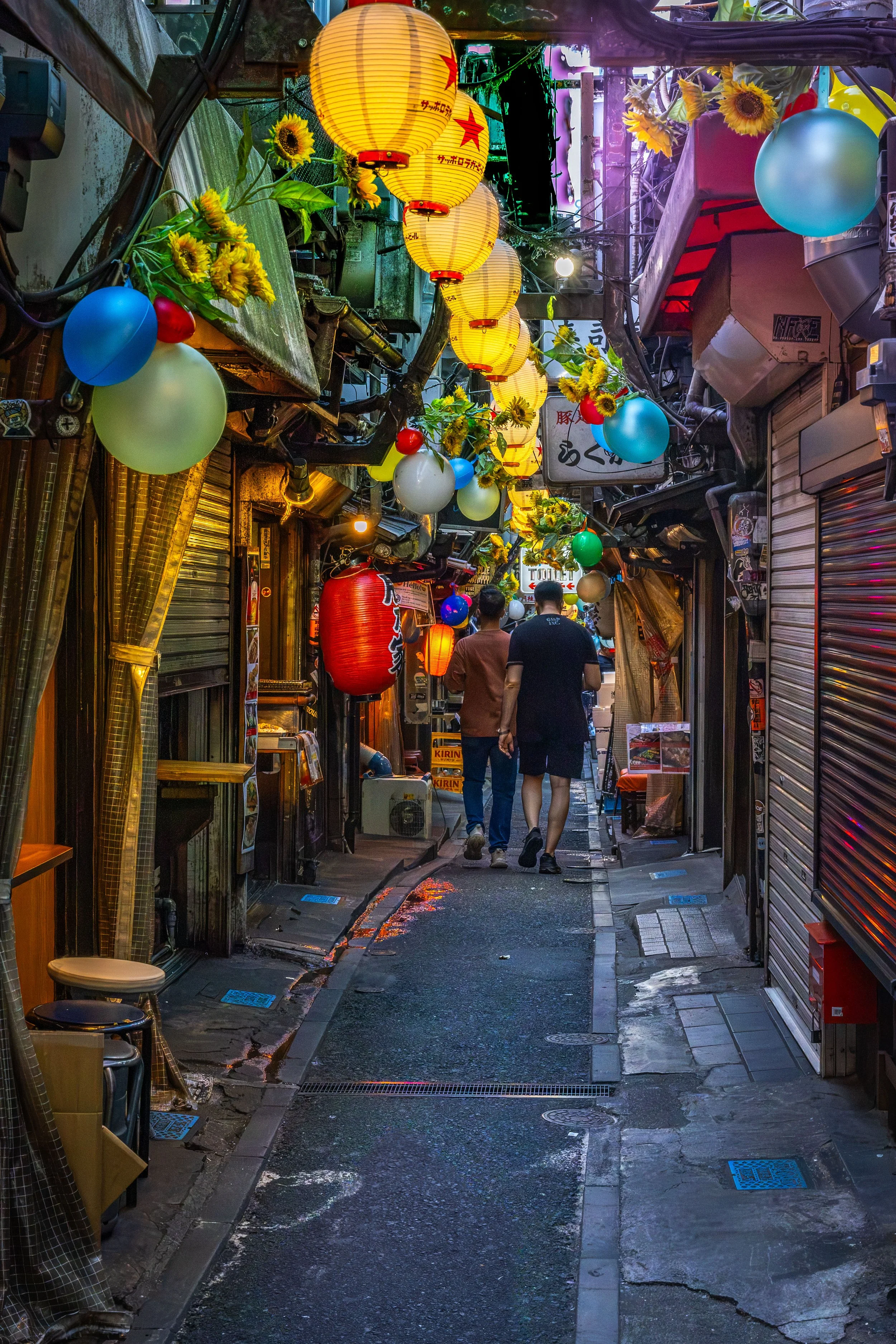 A narrow street in Japan decorated for a festival with paper lanterns, balloons, and sunflowers, featuring two men walking away from the camera.