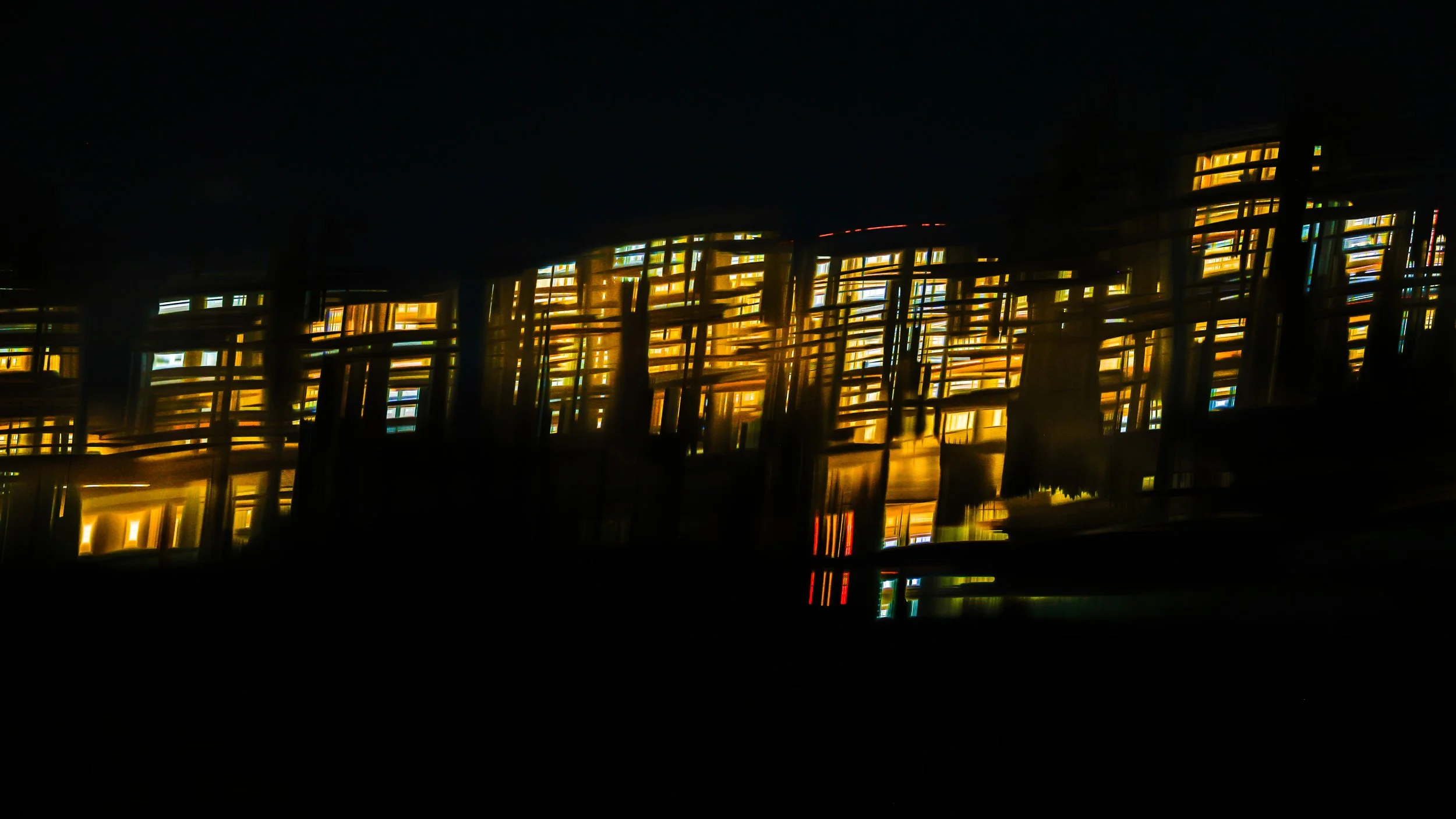 Night view of a modern building with illuminated windows and glass facade, reflecting colorful lights and dark sky.