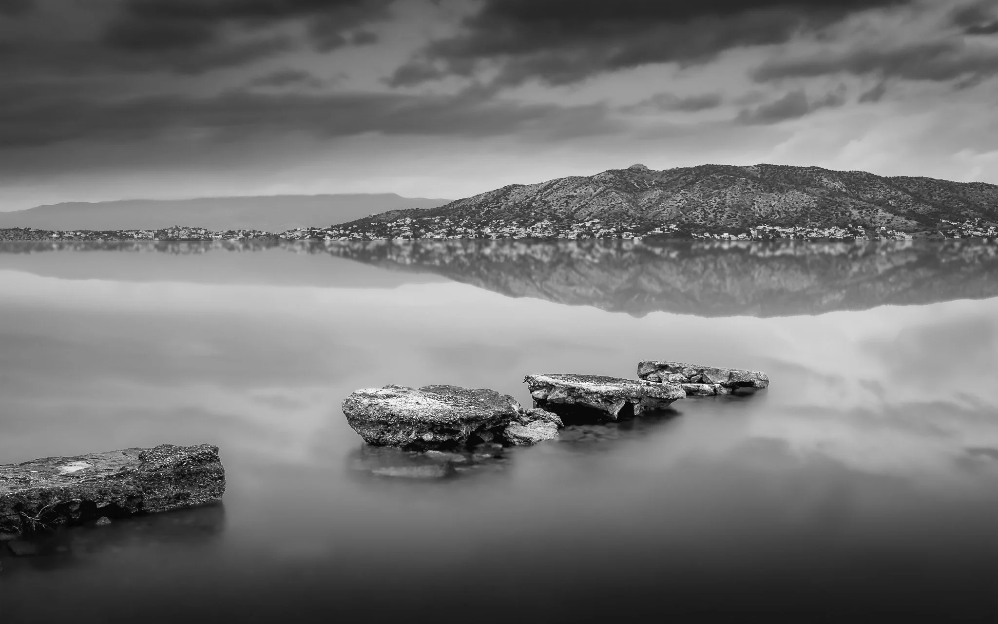 Black and white landscape photograph depicting rocky shoreline in the foreground, a calm lake reflecting the distant mountain range and buildings, with a cloudy sky overhead.