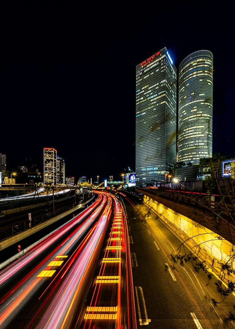 Nighttime cityscape with tall lit buildings and busy highway with red and white light trails from moving vehicles.