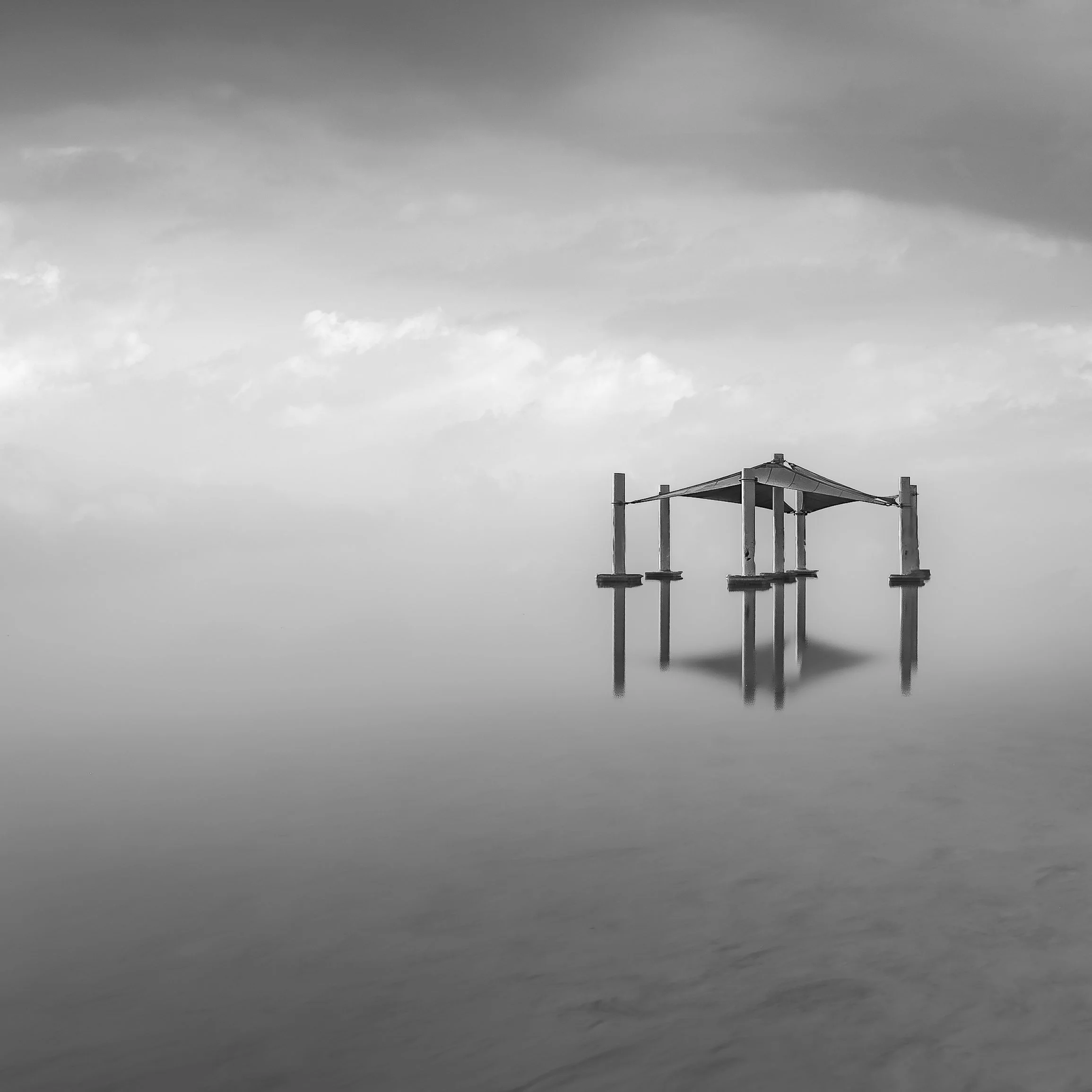 A black and white photo of a wooden pier with a canopy on calm water, reflecting the structure below the cloudy sky.