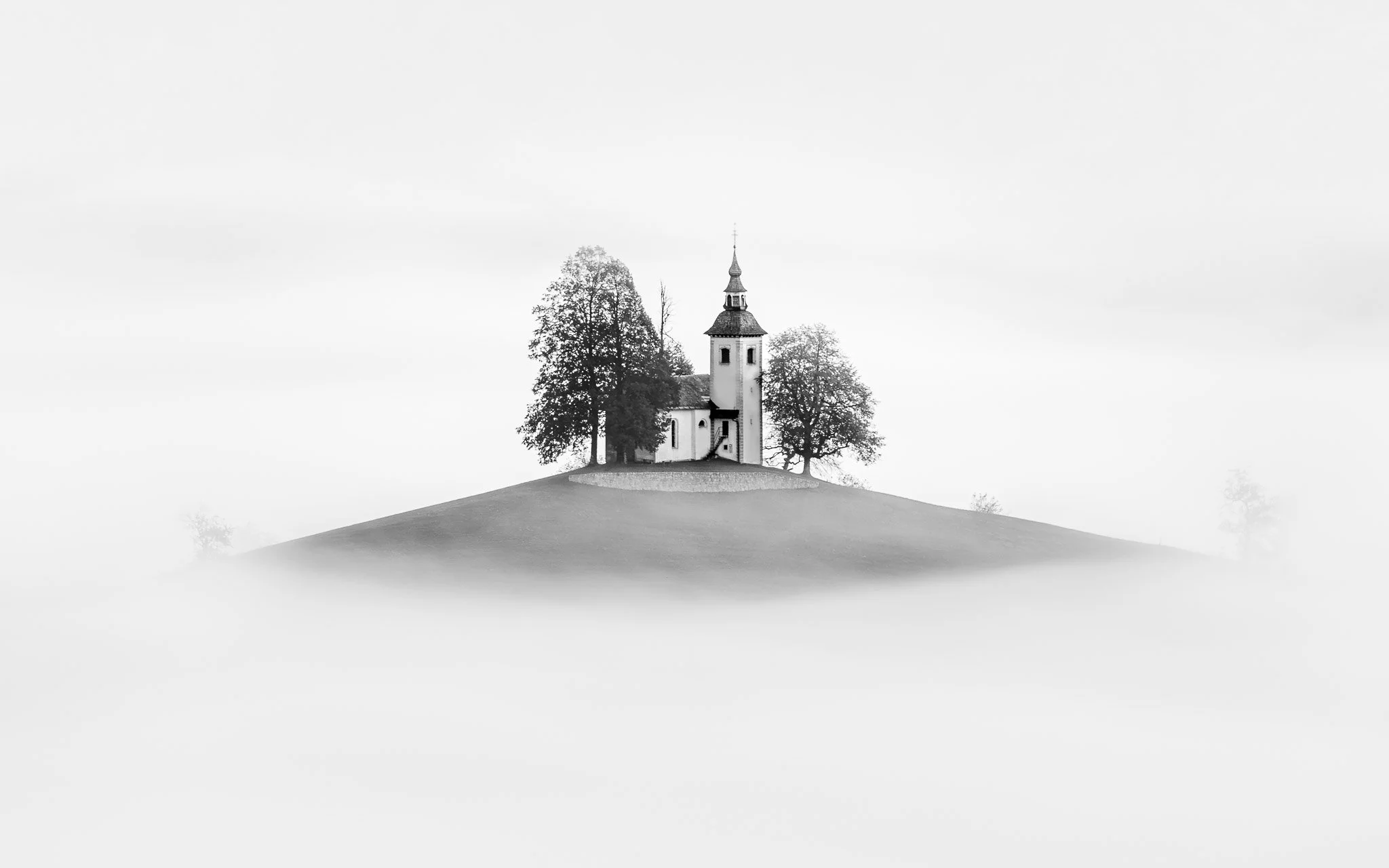 Minimalist black and white photograph of the Church of St. Thomas on a small hill, framed by trees and surrounded by dense white fog in Slovenia.