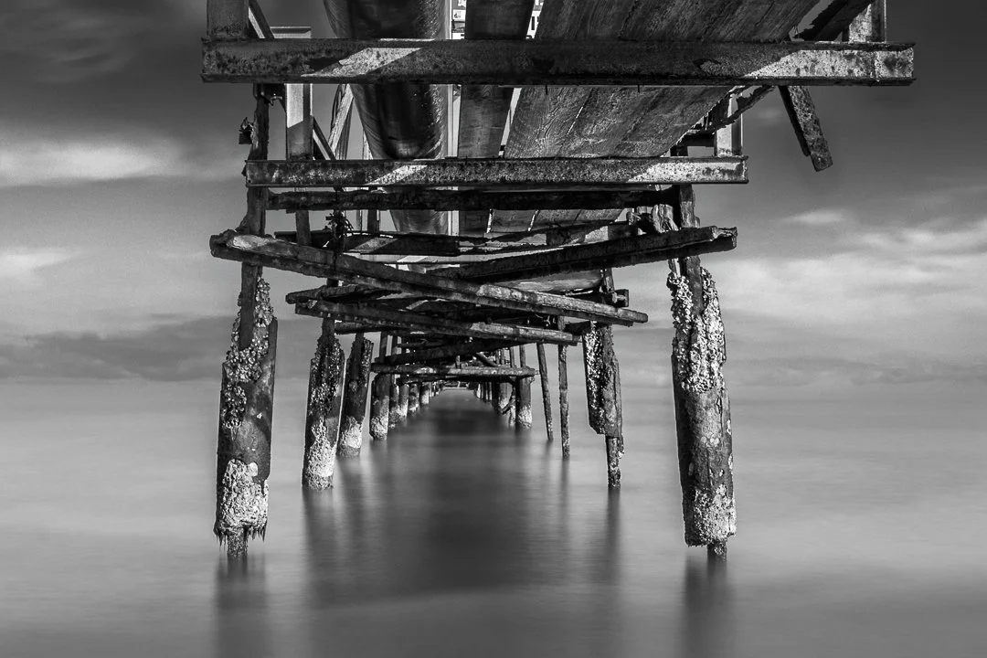 Black and white photo of a worn, wooden pier extending into the water with visible barnacle growth on the support beams.