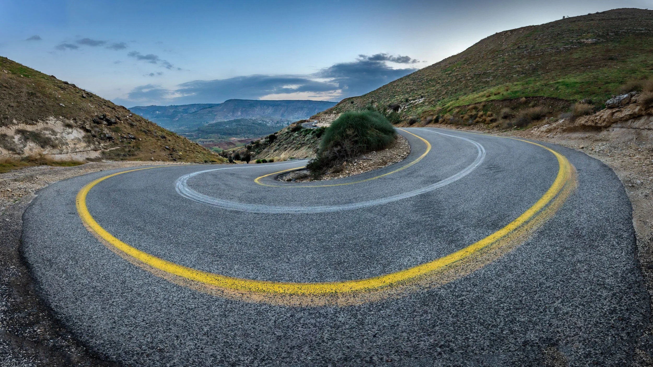 A winding mountain road with double yellow lines and guardrails on the side, surrounded by hills and cloudy sky.