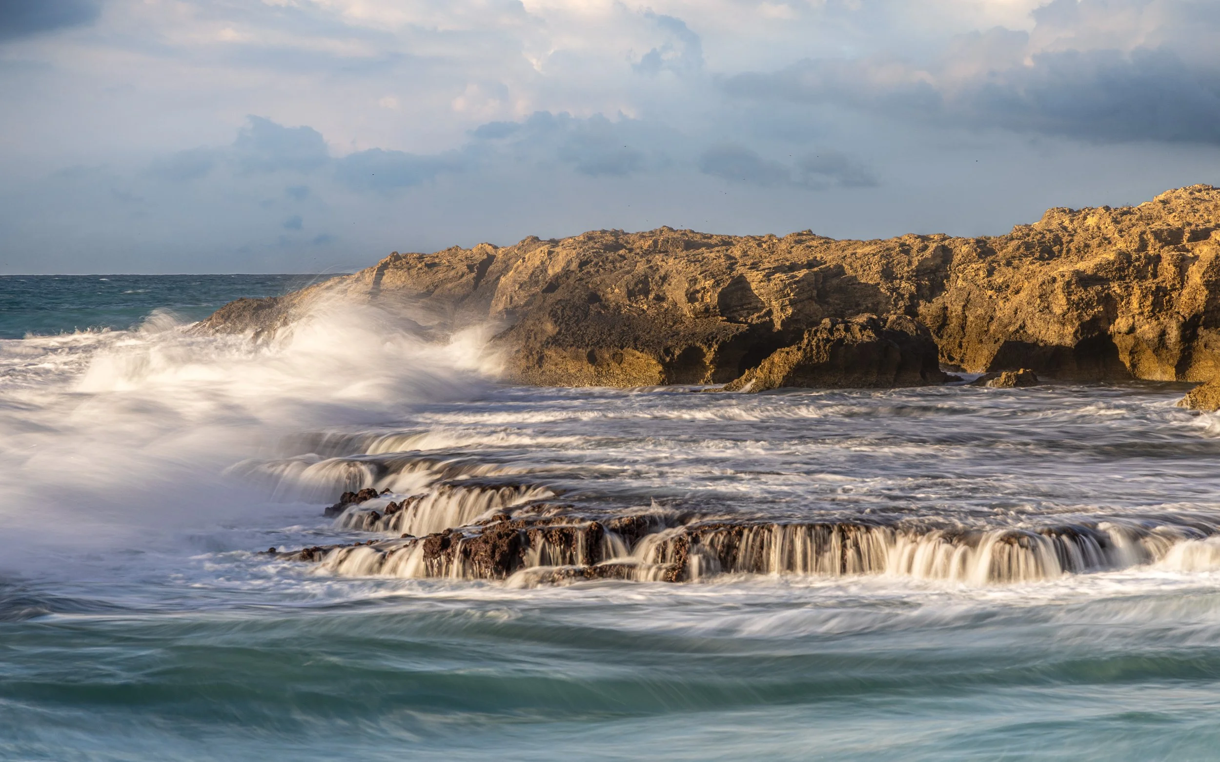 Waves crashing against rocky shoreline with clouds in the sky.